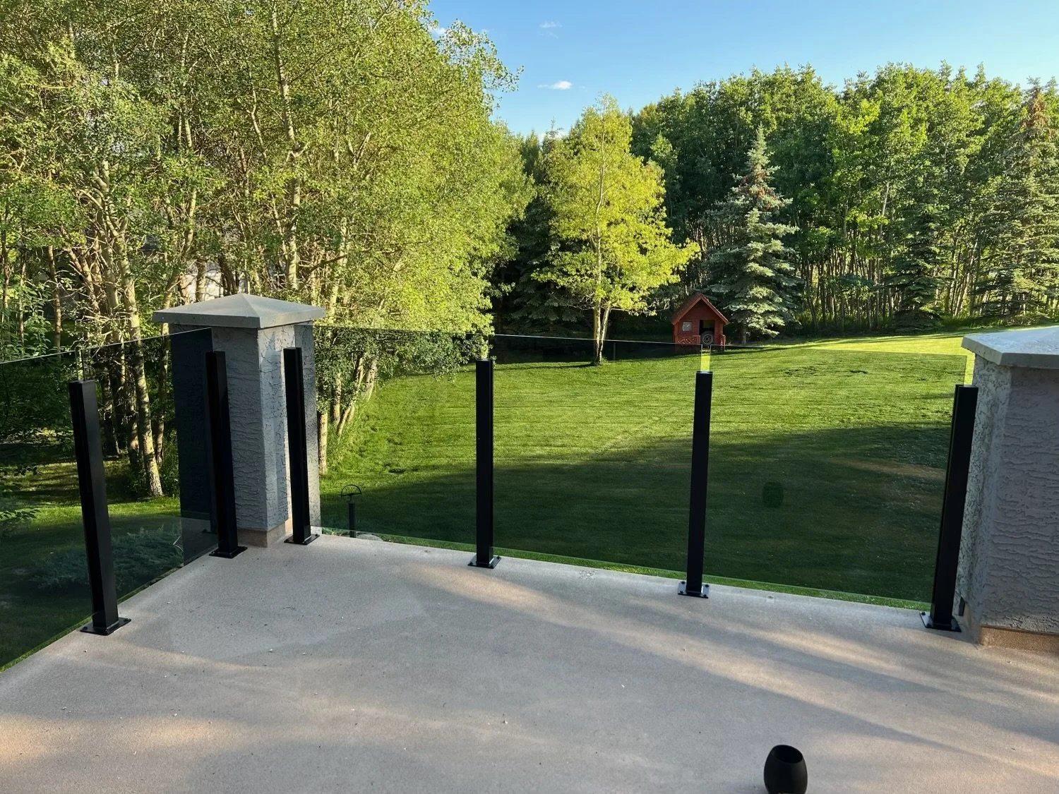 View from a patio with a glass railing, looking onto a grassy backyard with trees and a small red shed