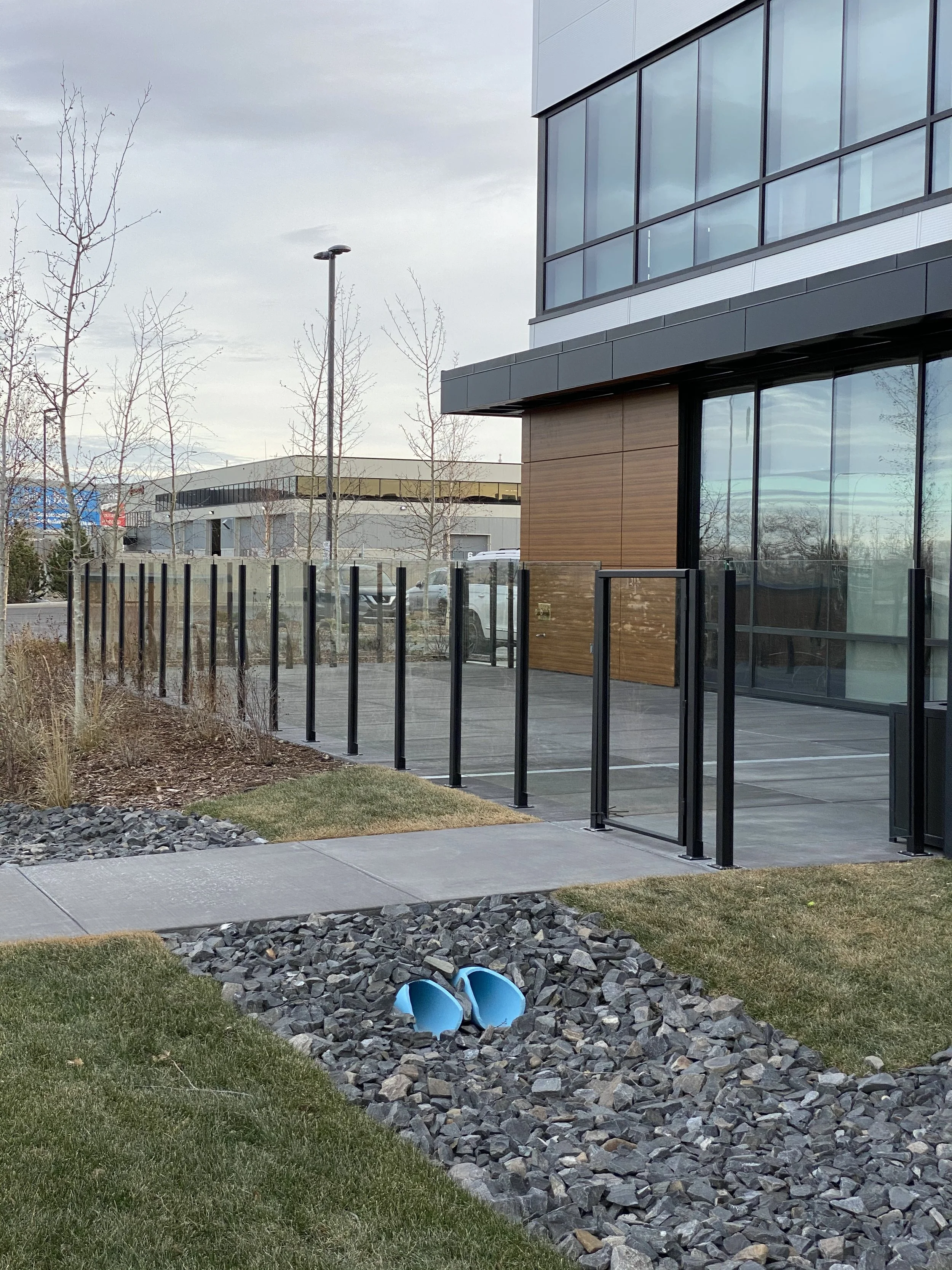 A modern building with large glass windows, a small patio area with a glass railing, and a landscaped yard with grass, rocks, and two blue slide pipes.