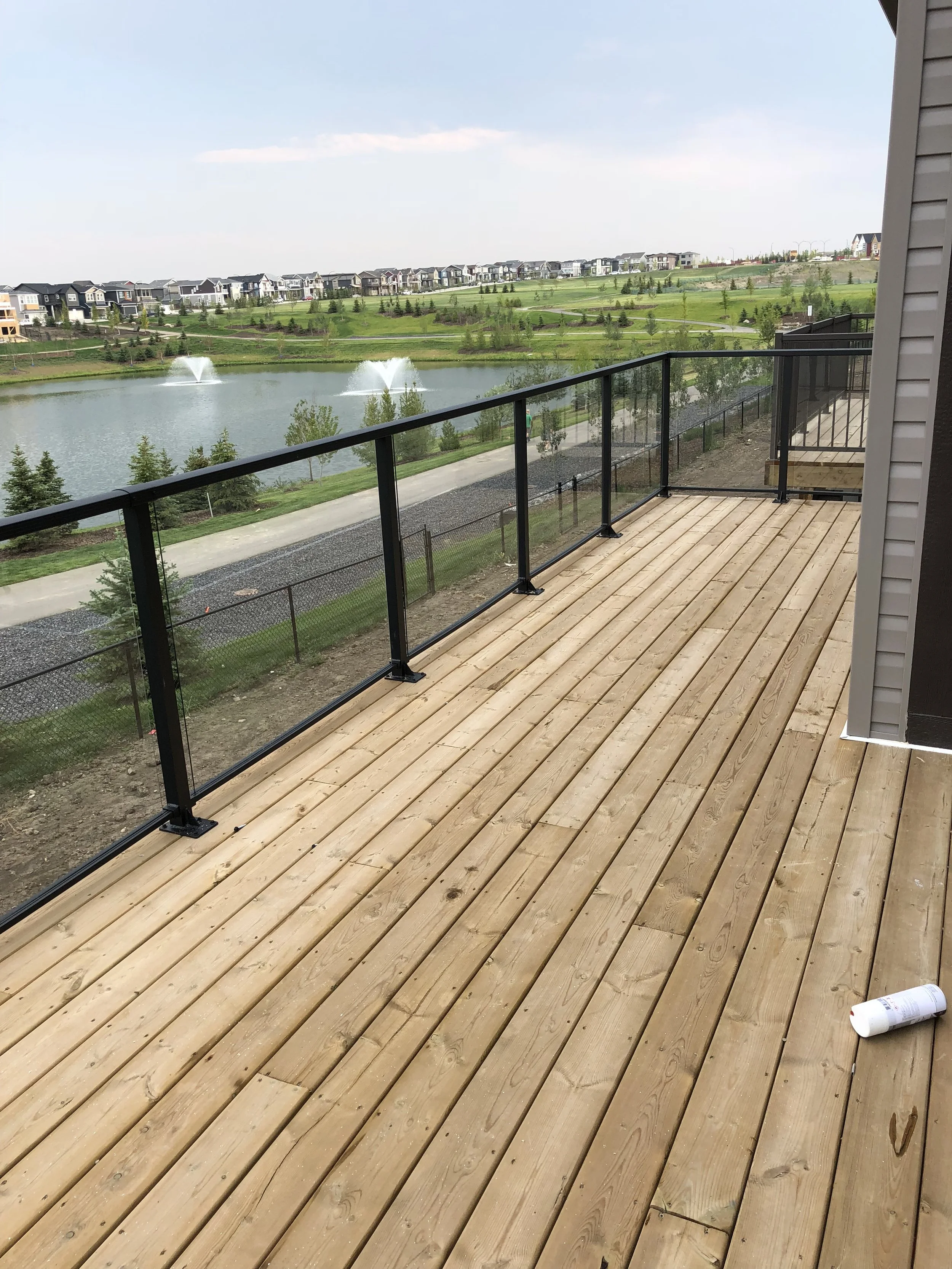 Wooden deck under construction with black metal railing and a scenic view of a pond with fountains, green grass, trees, and a row of houses in the background.