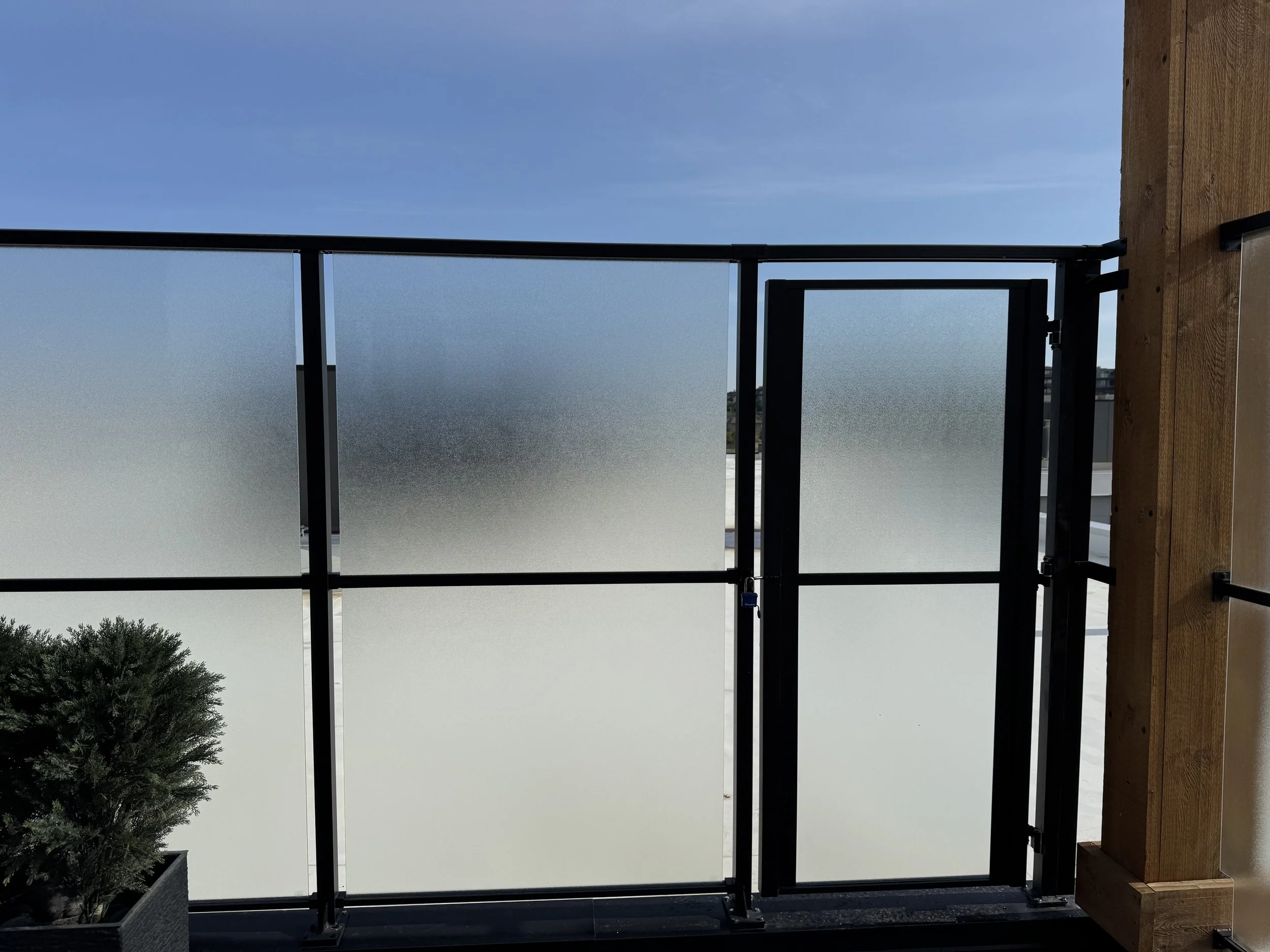 View of a balcony with a glass railing, a blue sky with some clouds, and a small tree in the lower left corner.