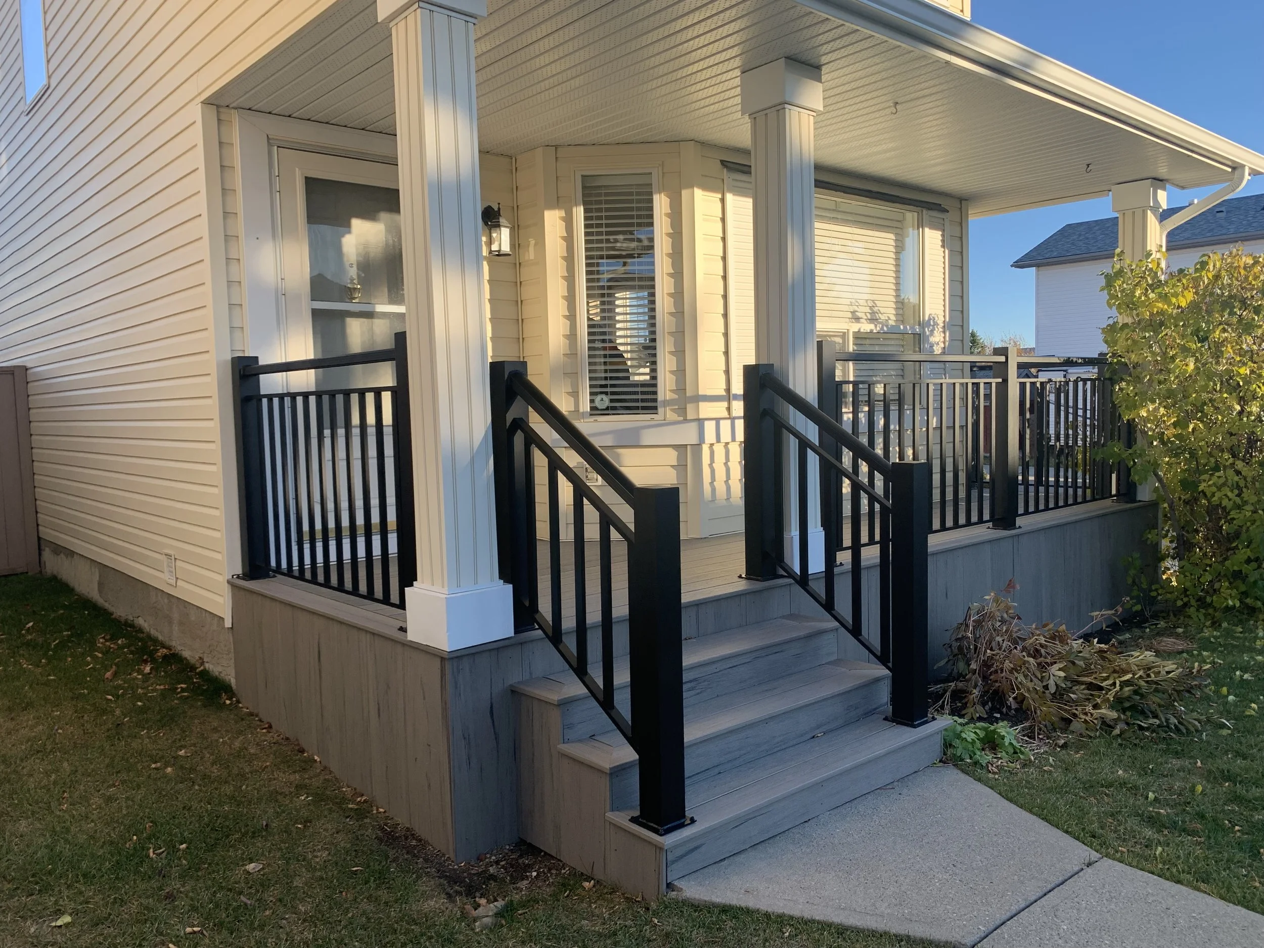 Front porch of a house with stairs, black railing, and a covered roof, beige siding, a window with blinds, a door with a screen, surrounded by greenery and a clear blue sky.