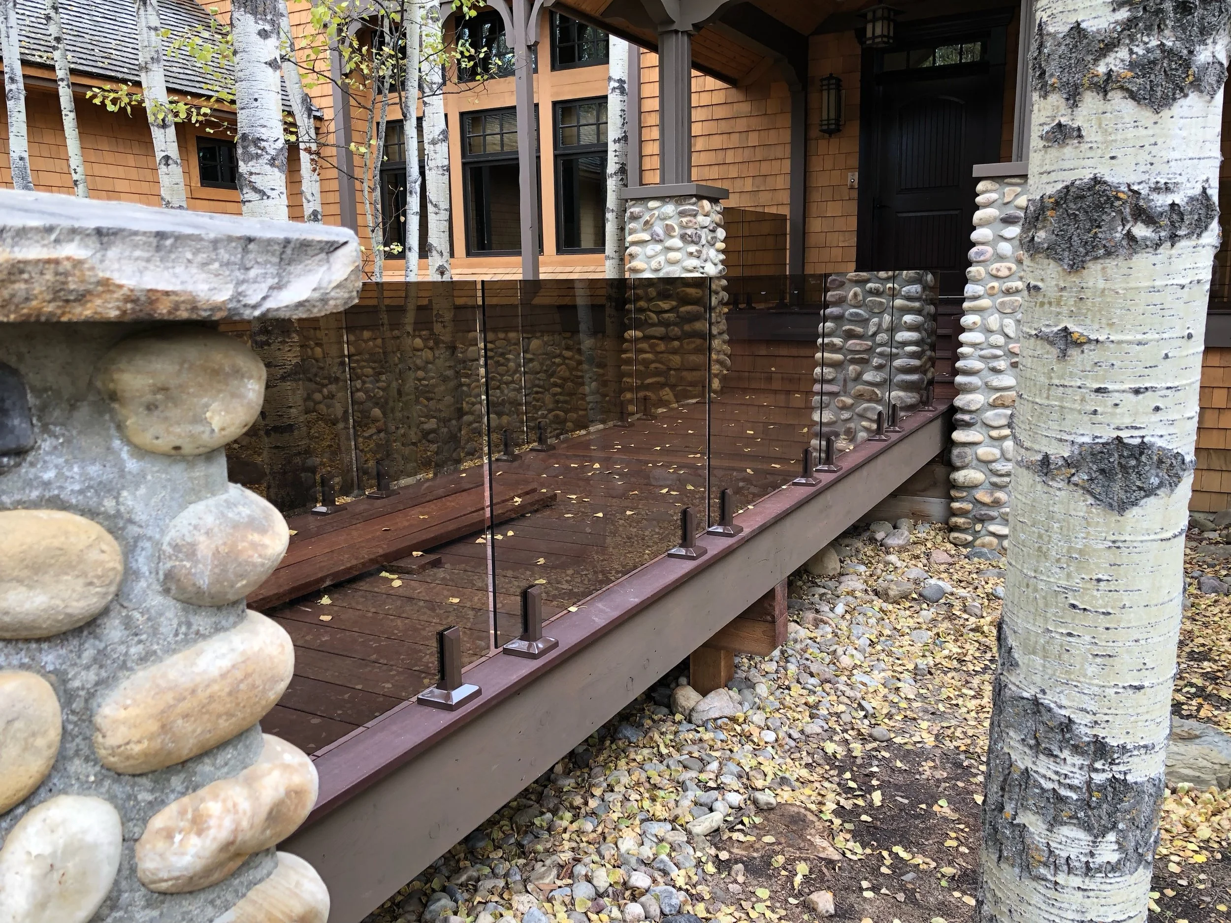 Close-up of a stone and wood railing in front of a house with wooden siding and multiple windows, with trees and autumn leaves on the ground.