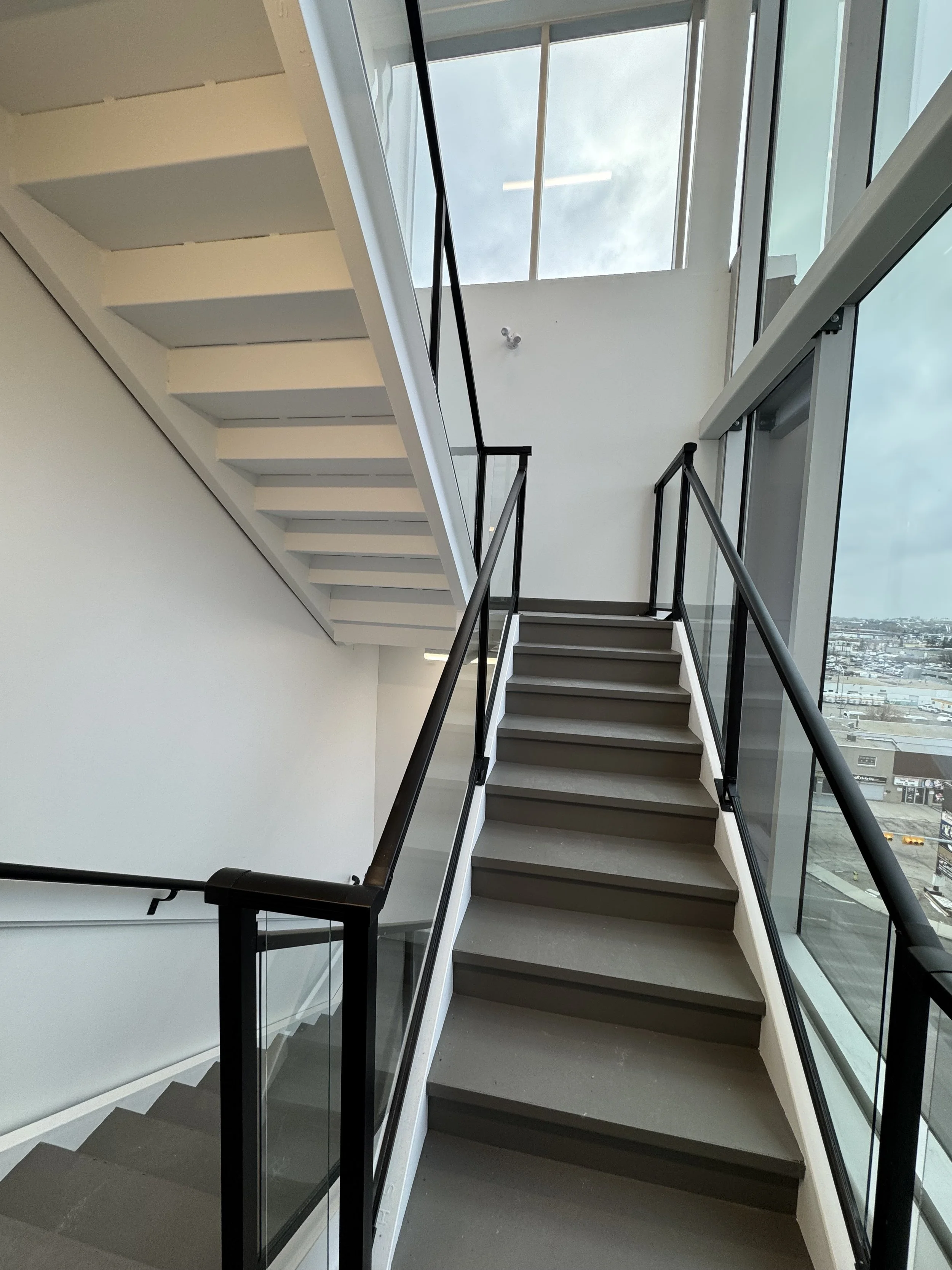 The image shows a modern stairwell with dark-colored steps and a sleek black metal and glass railing, with large windows allowing natural light and a view of the cityscape outside.