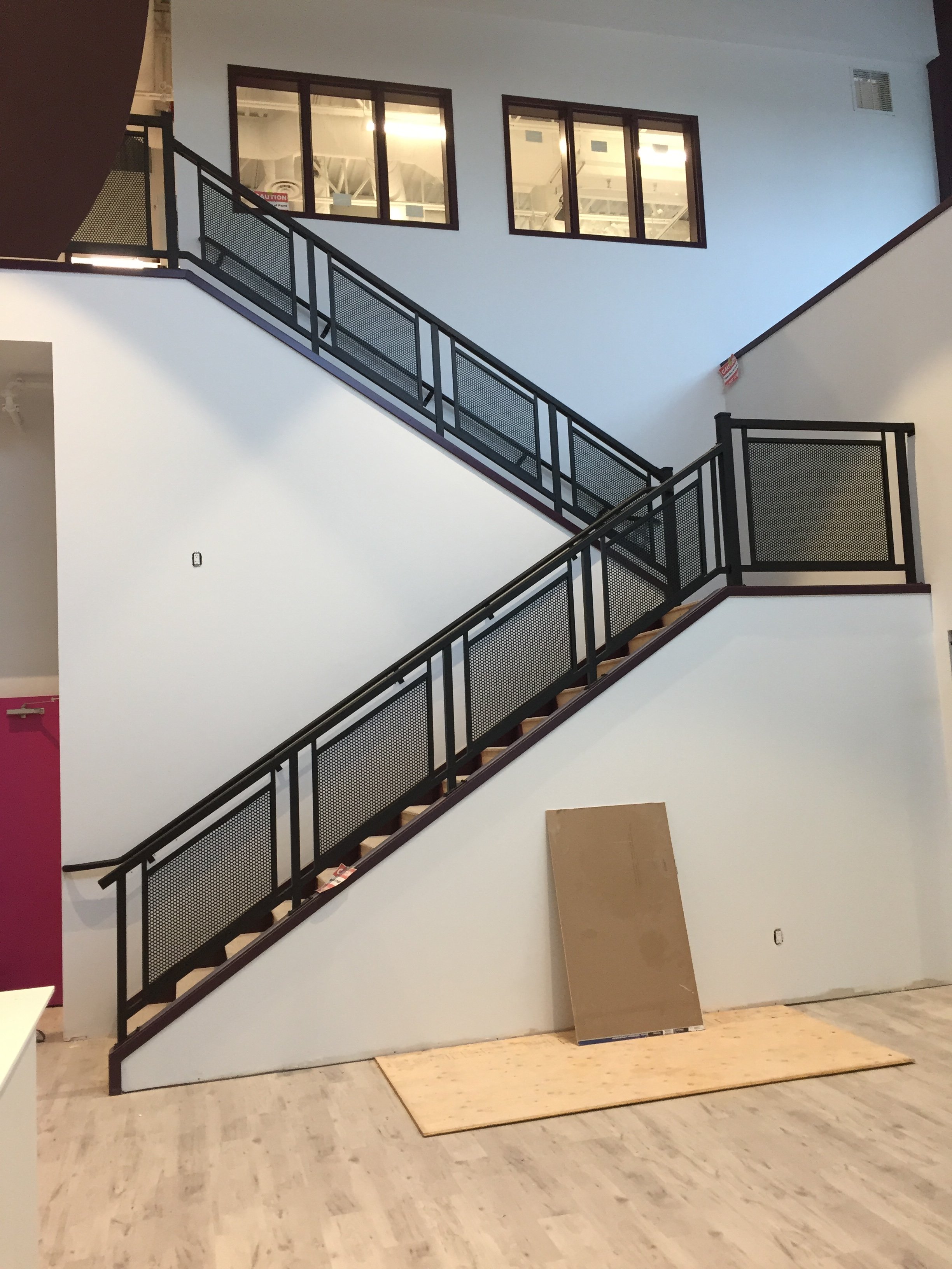 Interior of a building under construction with a staircase and black metal railing, a carpet roll, and a plywood board on the floor.