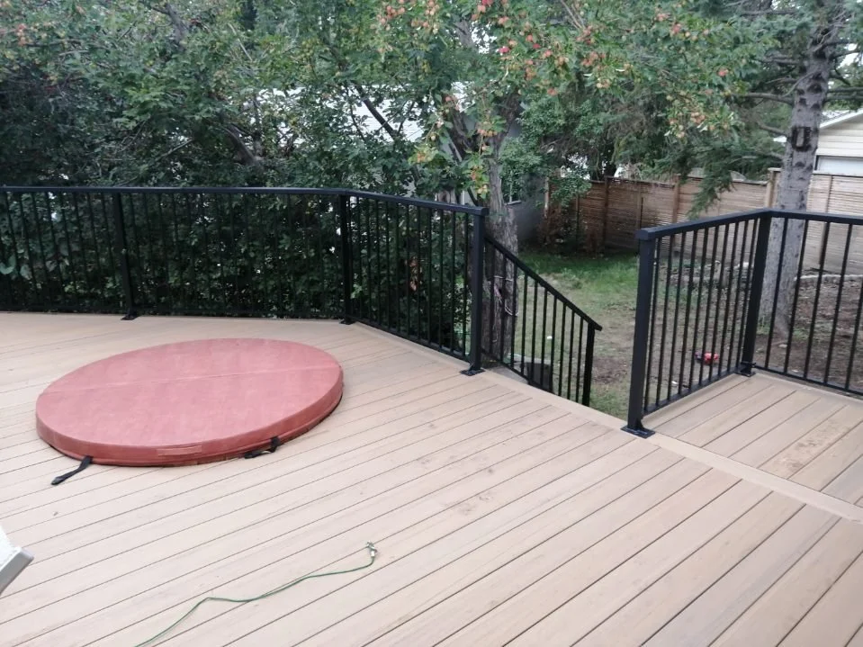 A wooden deck with a black metal railing, a red circular hot tub cover, and trees and grass in the backyard.