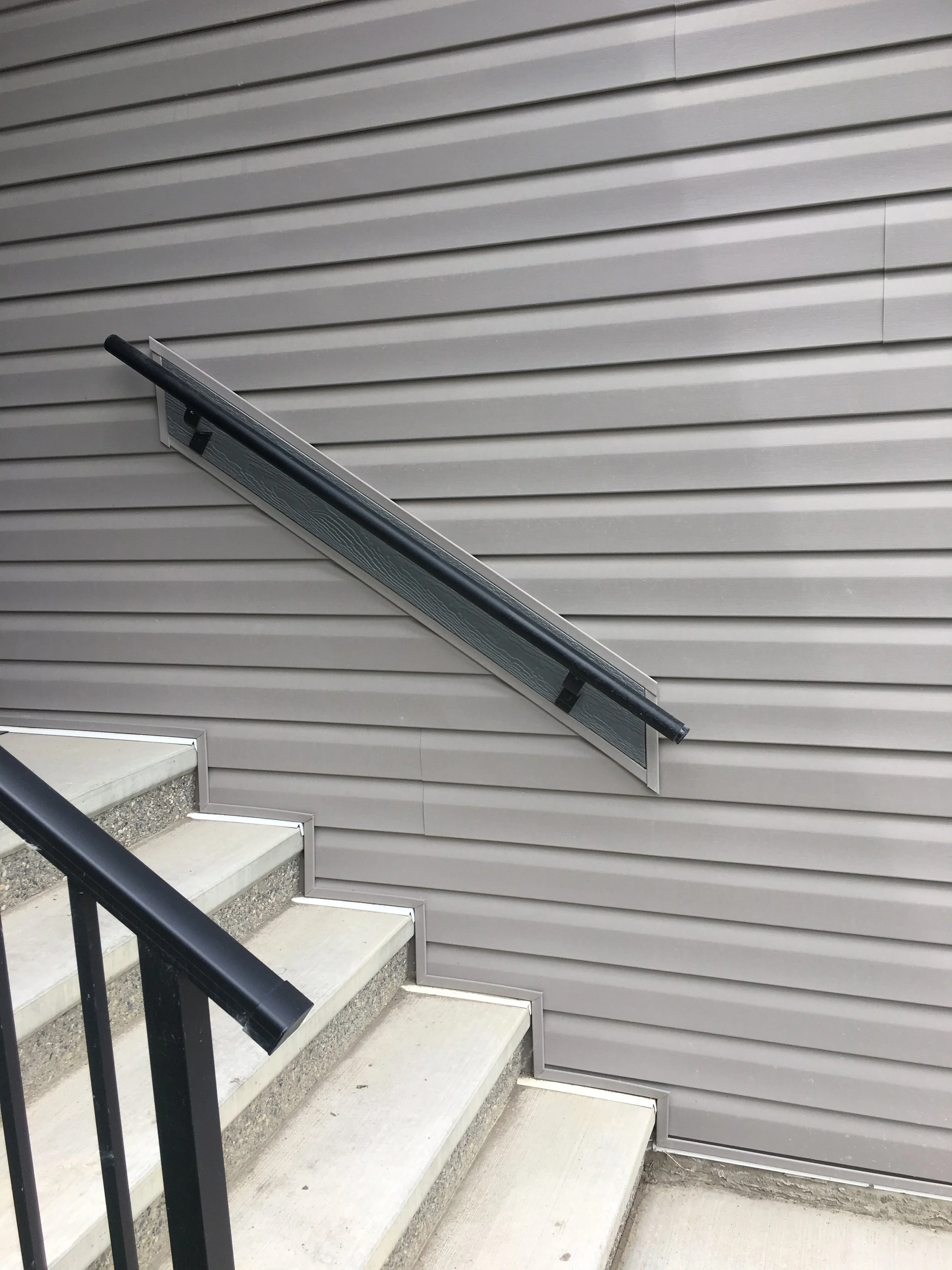 Outdoor staircase with concrete steps and black metal railing, adjacent to a house wall with horizontal gray siding.