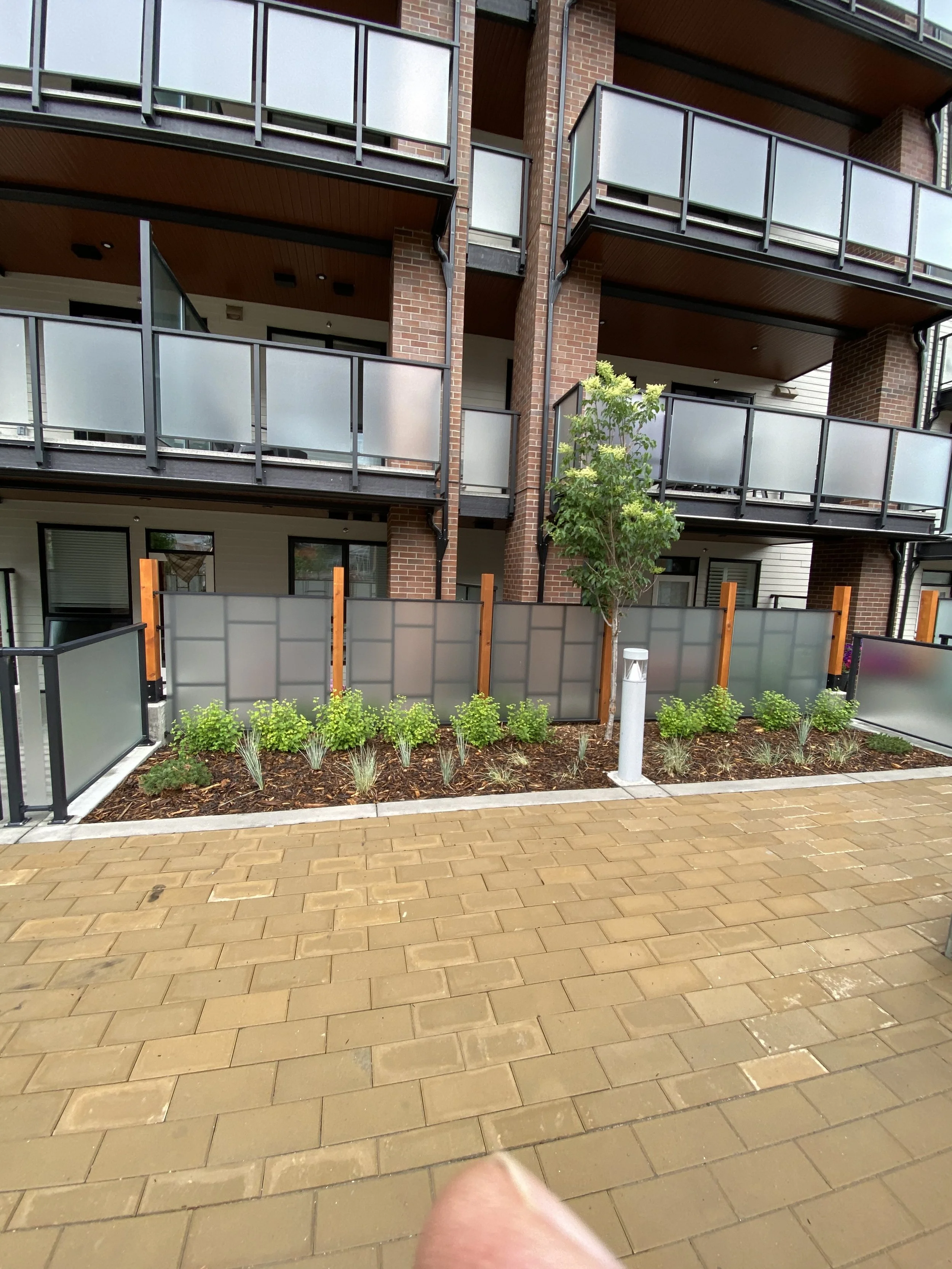 An apartment building with brown brick walls and multiple balconies with frosted glass railings, a small landscaped area with various green plants in front, and a paved walkway.