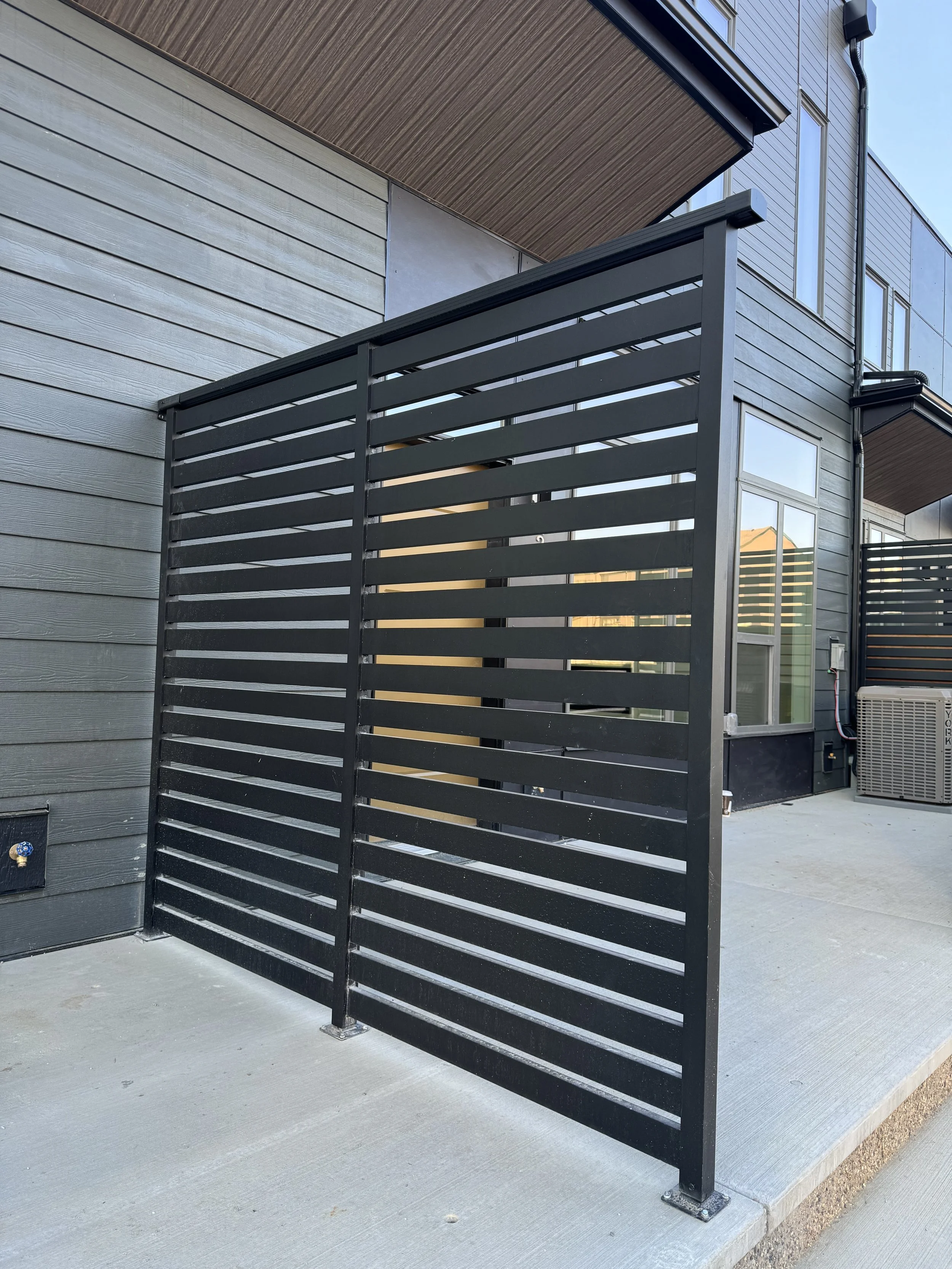 A black metal privacy screen with horizontal slats in front of a modern gray wooden exterior wall of a building with multiple windows. A concrete patio surface extends along the building.