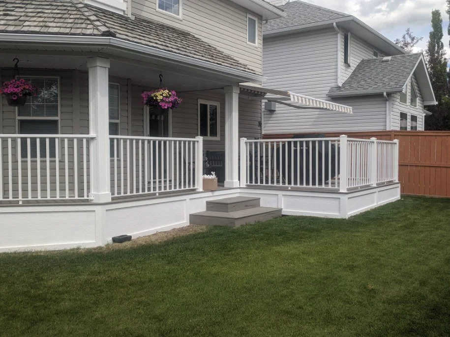 Backyard patio deck with white railing, grey steps, and a beige house with flower pots hanging on the wall.