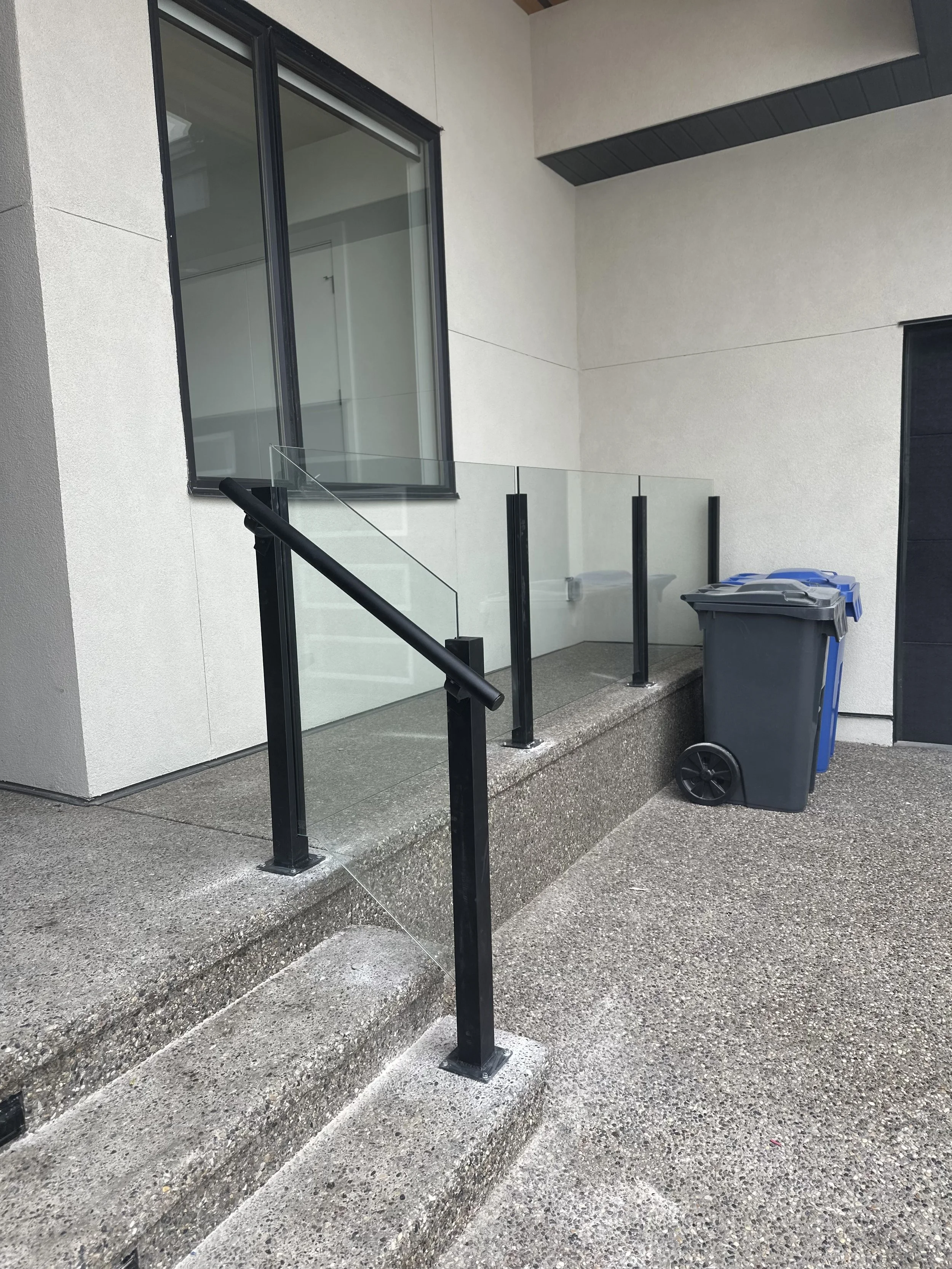 Exterior view of a modern residential building with a small porch, glass railing, a trash bin, and concrete steps.