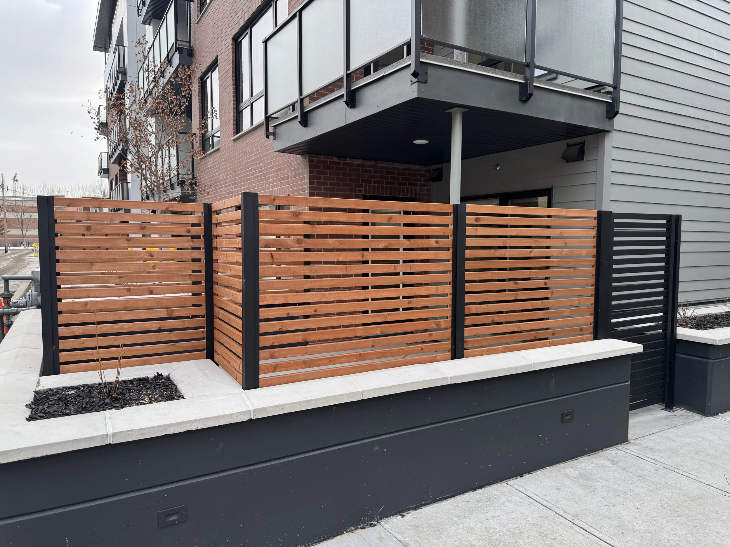 Modern apartment building with brick and gray siding, featuring balconies with glass and metal railings, and a wooden privacy fence in the foreground. There are small plant beds with mulch and a sidewalk in front.