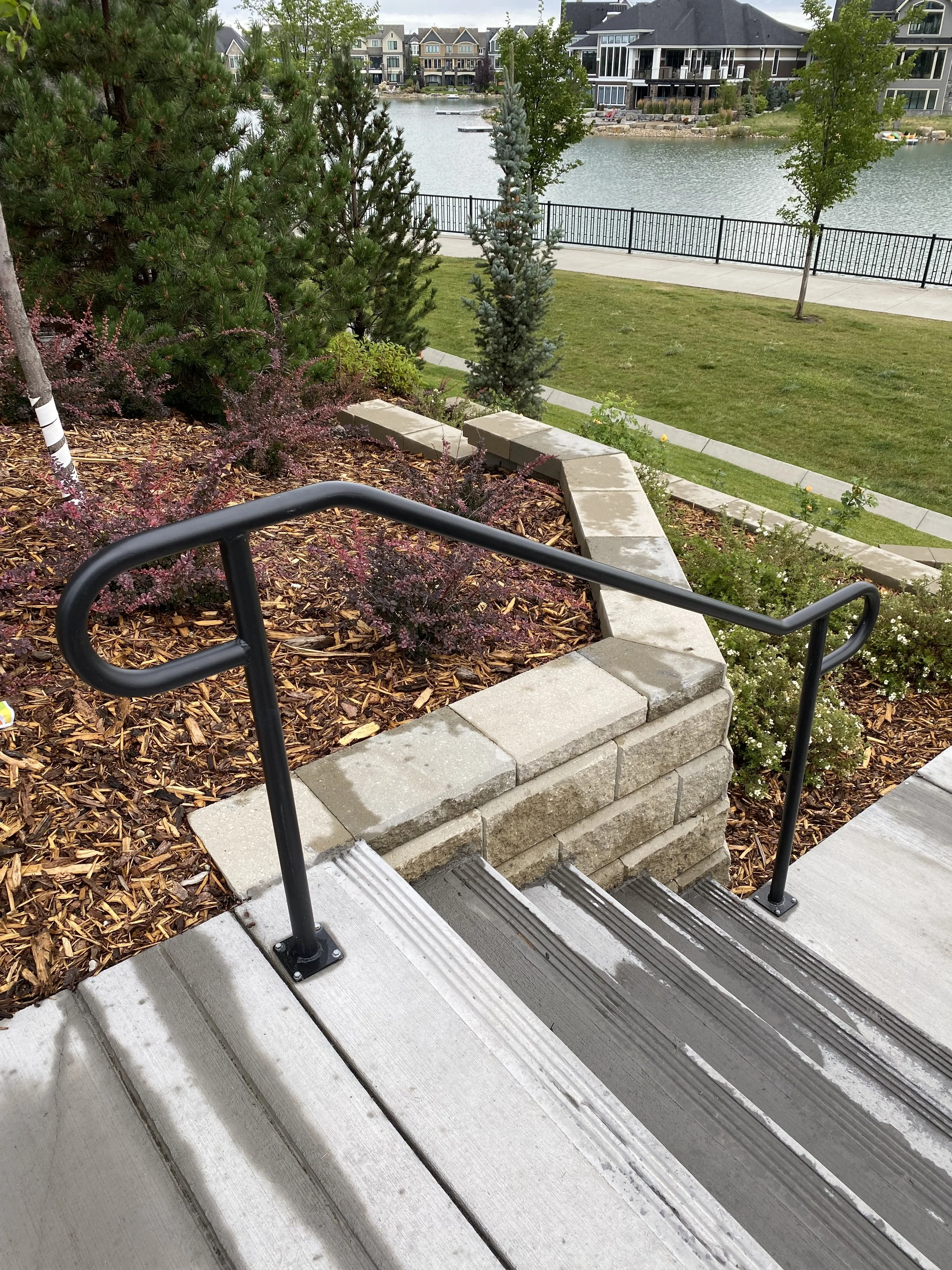 View of stairs with handrails leading down to a landscaped area with mulch, shrubs, and trees, overlooking a park or lake with houses and buildings in the background.