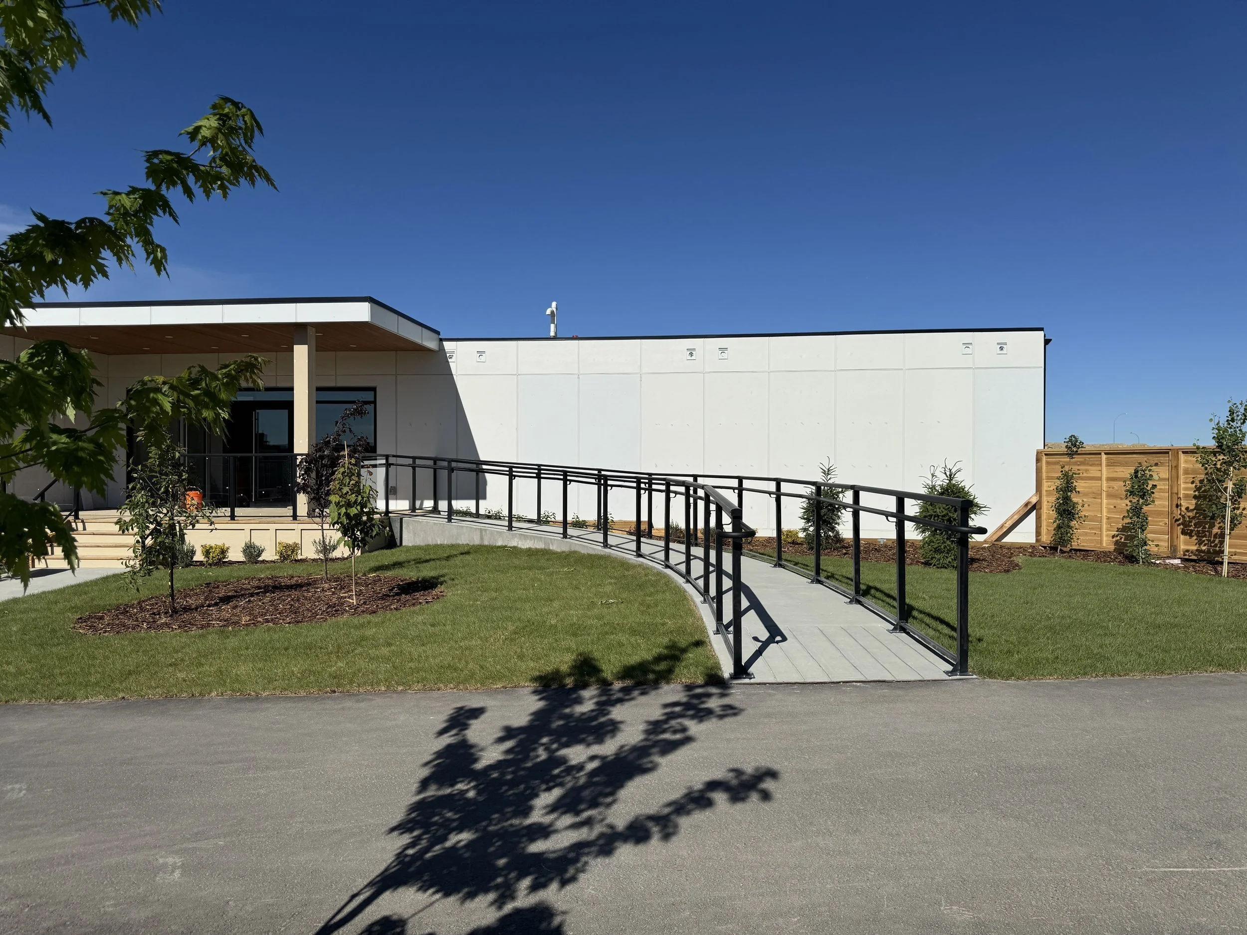 Modern building with white facade, large windows, and a curved ramp with black railing leading to an entrance, surrounded by a green lawn and trees under a clear blue sky.