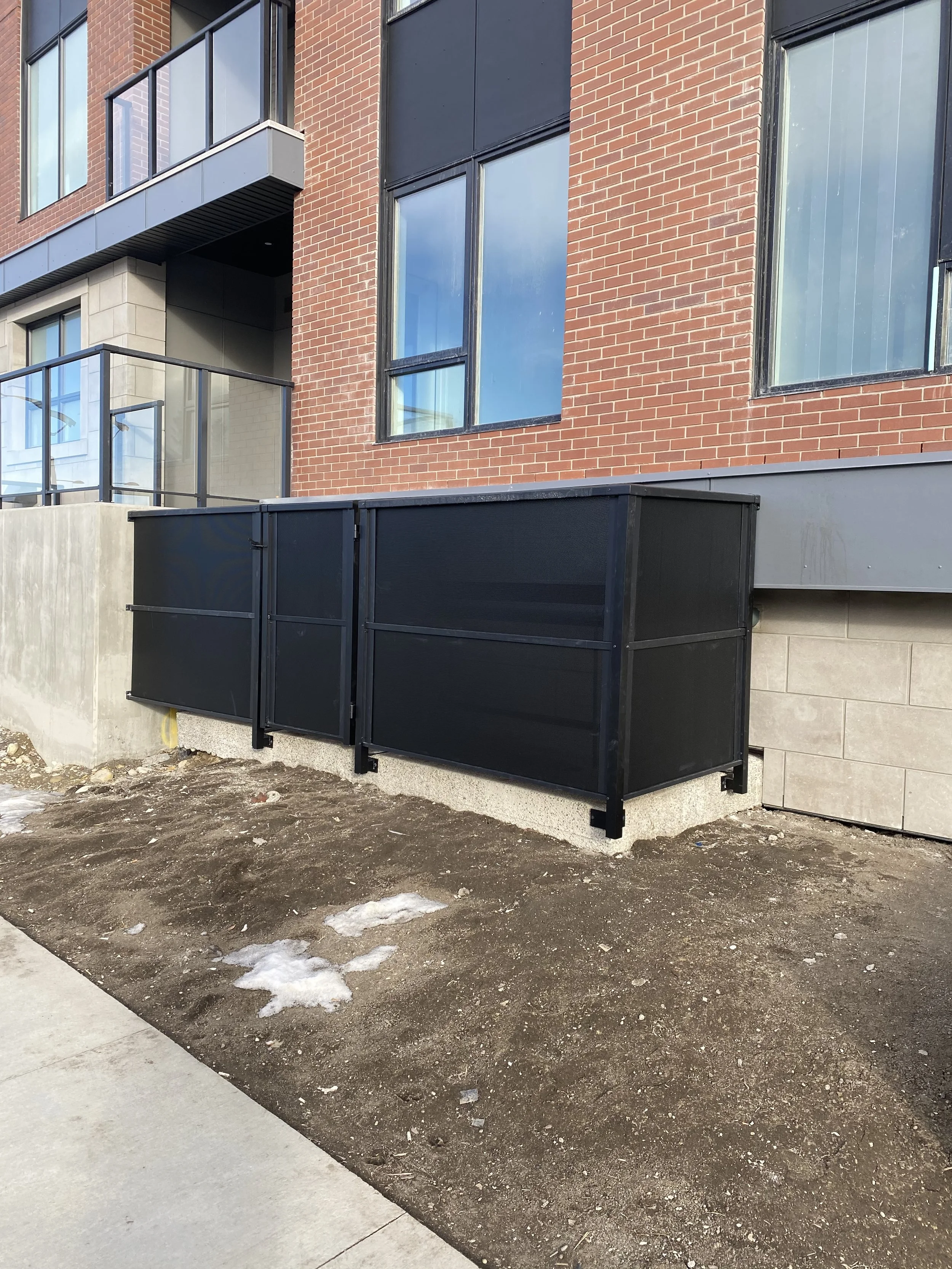 Exterior view of a modern apartment building with brick walls, large windows, and black metal railings on the balconies. In the foreground, there is a patch of dirt with some small patches of snow, and a concrete sidewalk.