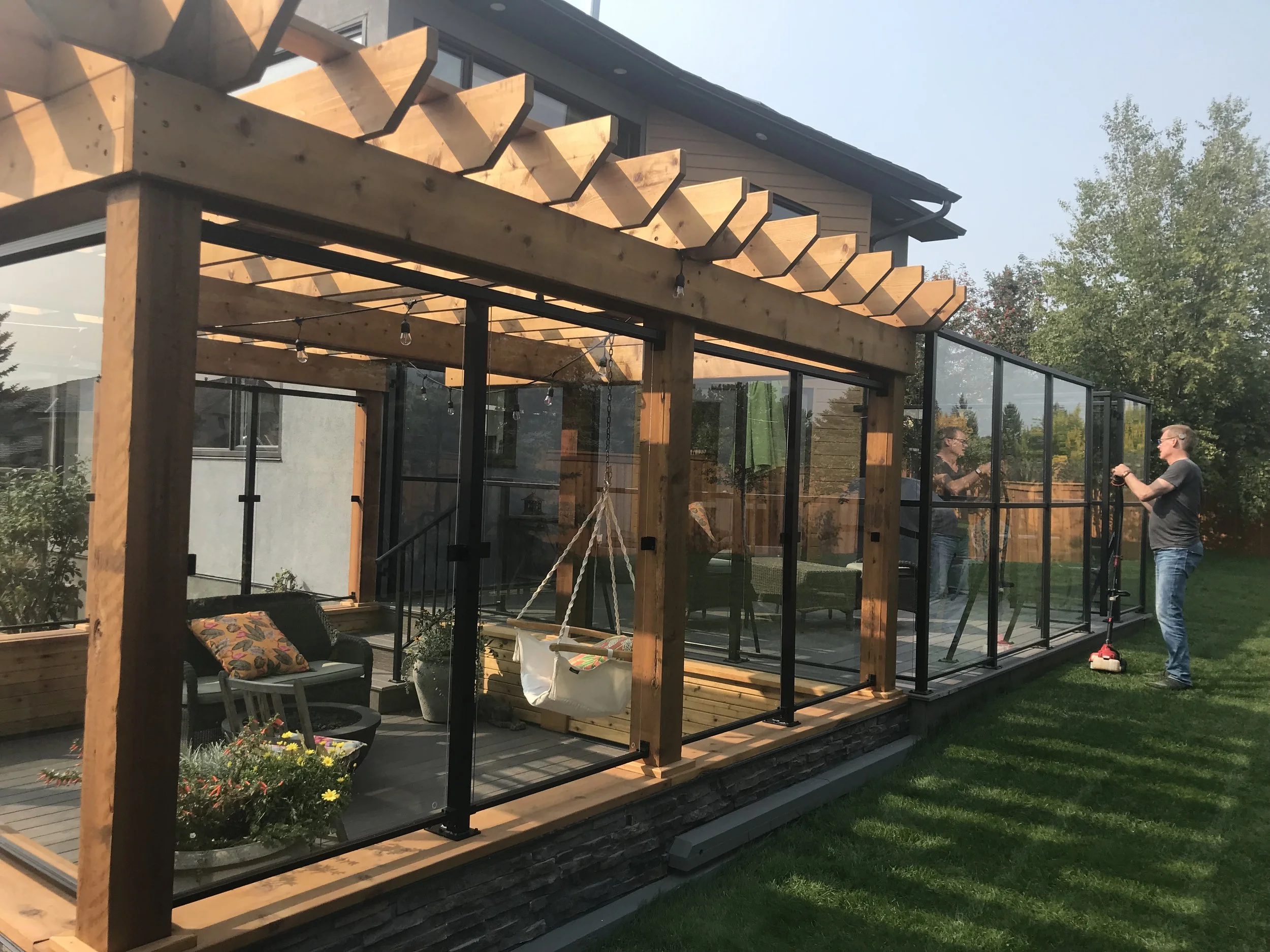 A backyard patio with a wooden pergola under construction, two men standing nearby, one holding a tool, and glass walls surrounding the area.