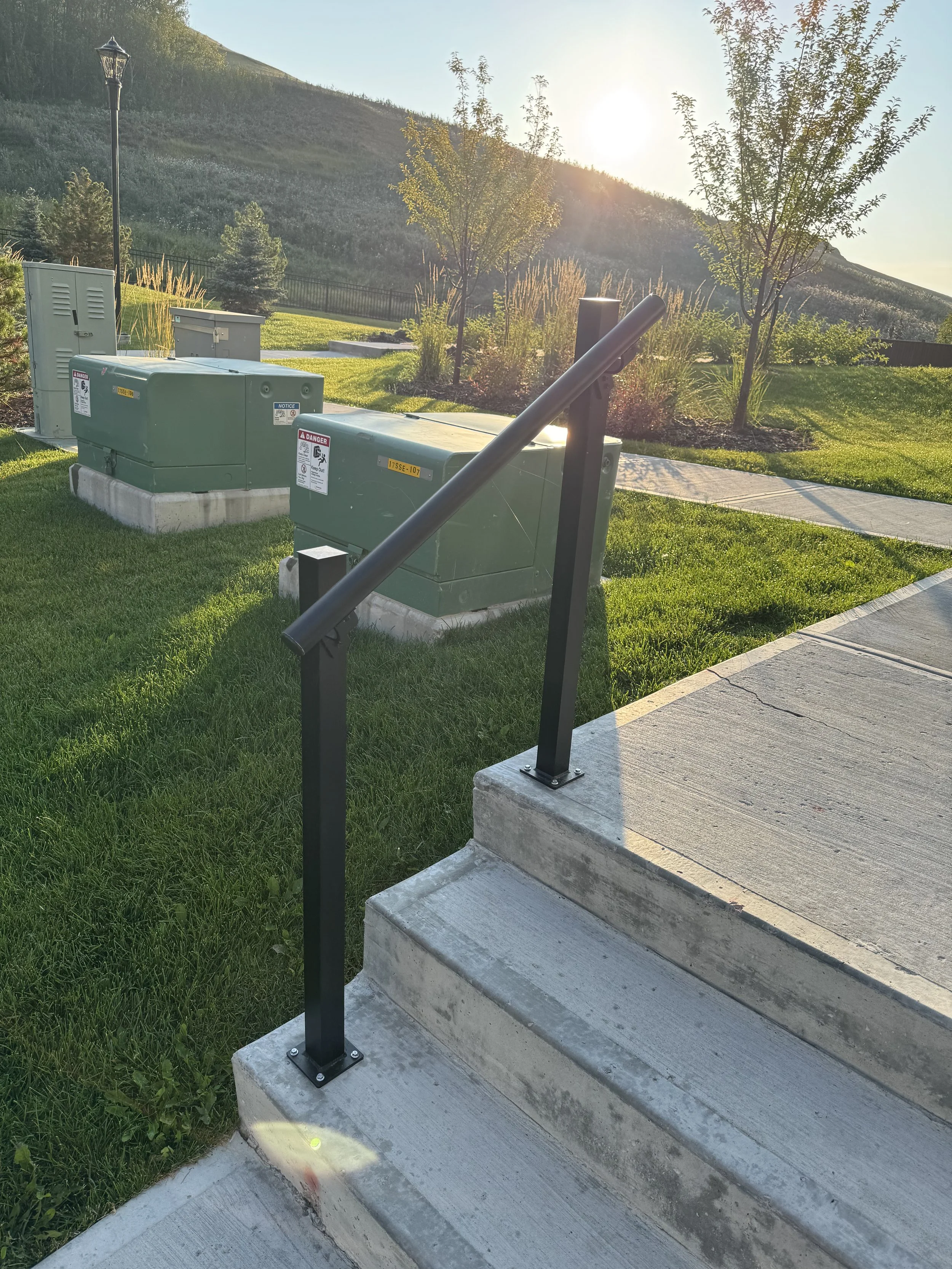 Electrical boxes and a handrail next to concrete stairs on a sidewalk, with a grassy lawn, trees, a fence, and a hillside in the background during sunset.