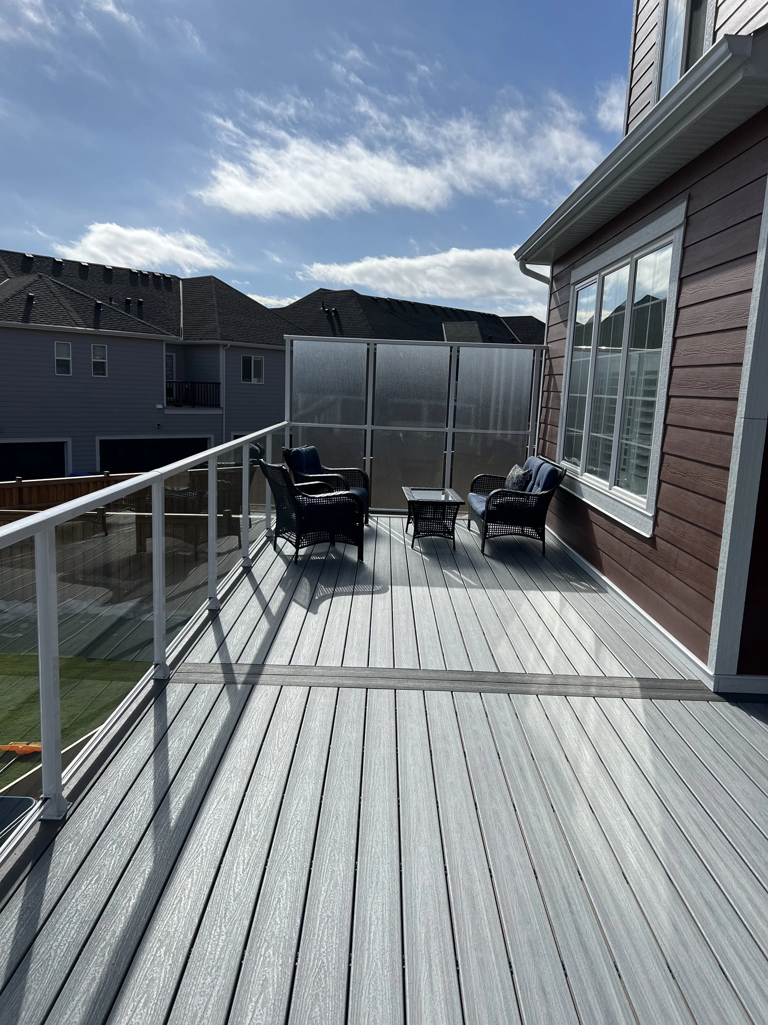 A backyard deck with outdoor furniture and a privacy screen, adjacent to a house with red horizontal siding, under a blue sky with scattered clouds.