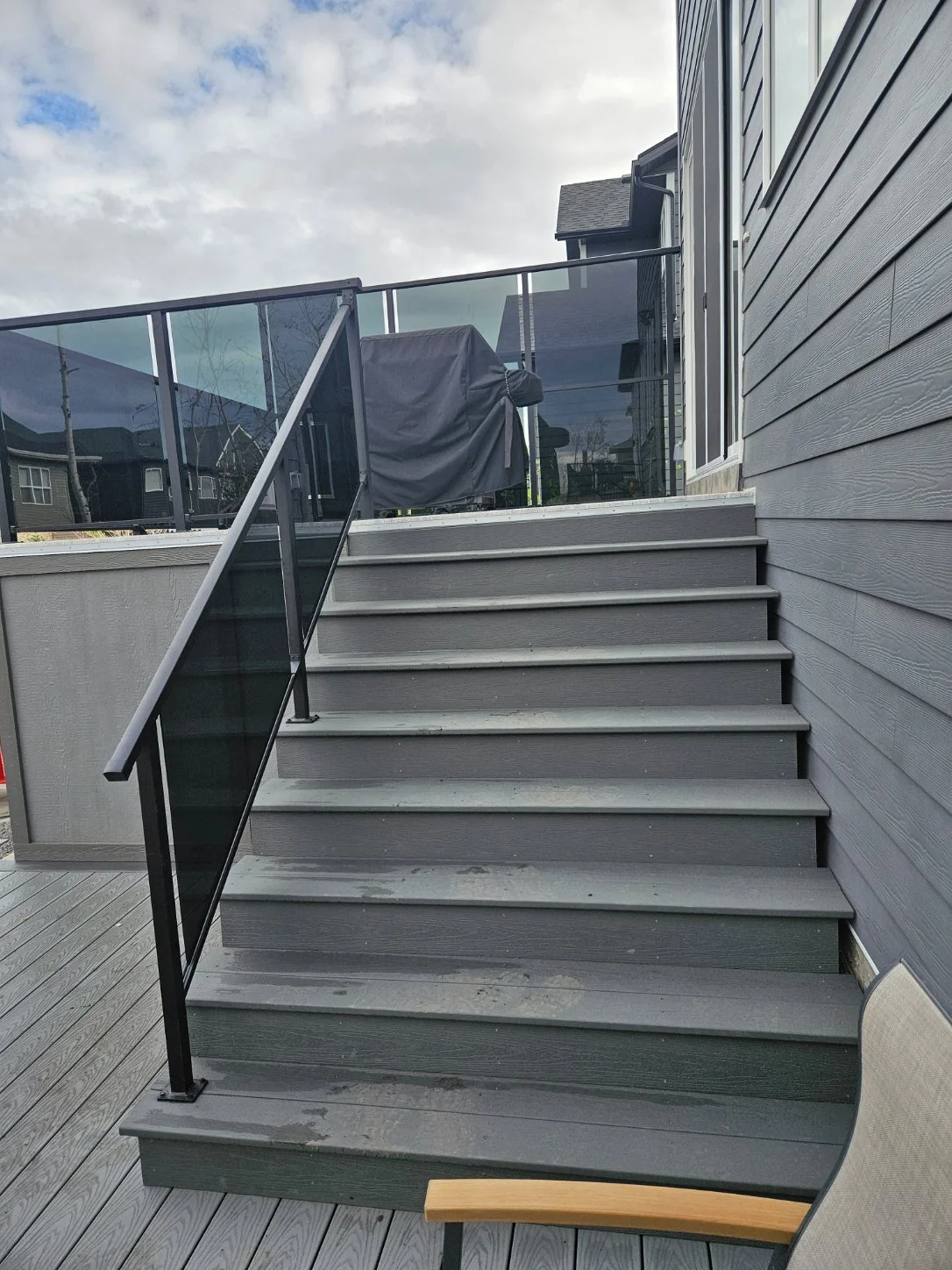 Gray wooden deck stairs with black metal and glass railing, a chair with light-colored wooden armrest, and a covered grill on the upper deck. Residential houses and cloudy sky in the background.