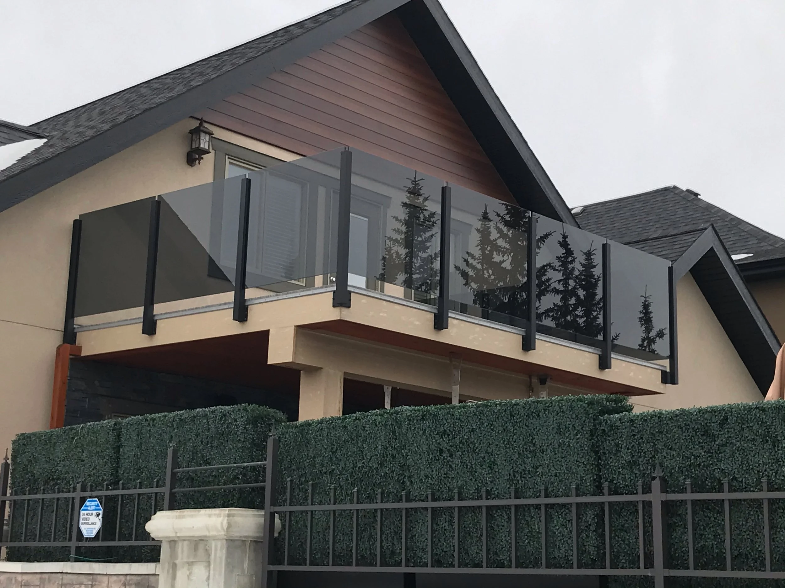 A modern multi-story house with a balcony surrounded by a metal fence and tall hedges. The house has beige and reddish-brown exterior walls, a black roof, and large windows. There is a lamp attached to the house exterior near the balcony.