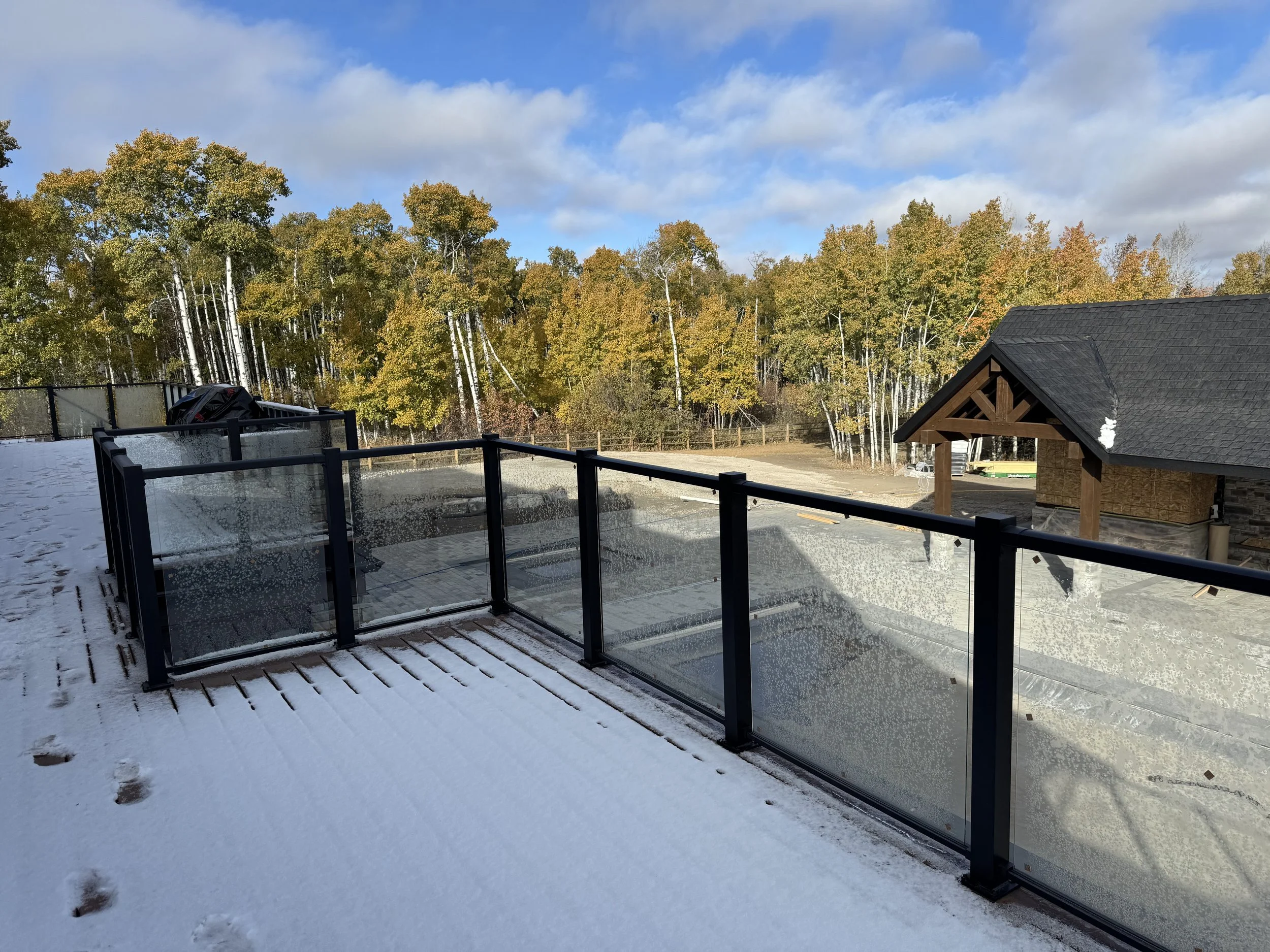 Snow-covered deck with footprints, glass railing, snow-covered landscape, wooden building, and a forest of trees with yellow and green leaves under a partly cloudy blue sky.