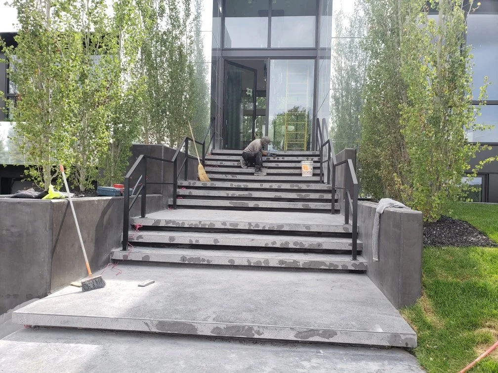 Person working on outdoor concrete stairs leading to a glass building entrance, with tools and construction materials around.