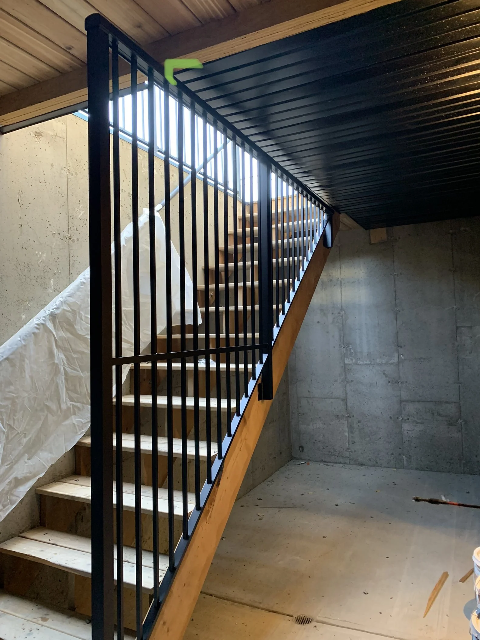 Interior view of a staircase under construction with wooden steps, a black metal railing, and an open space with a vent on the concrete floor.