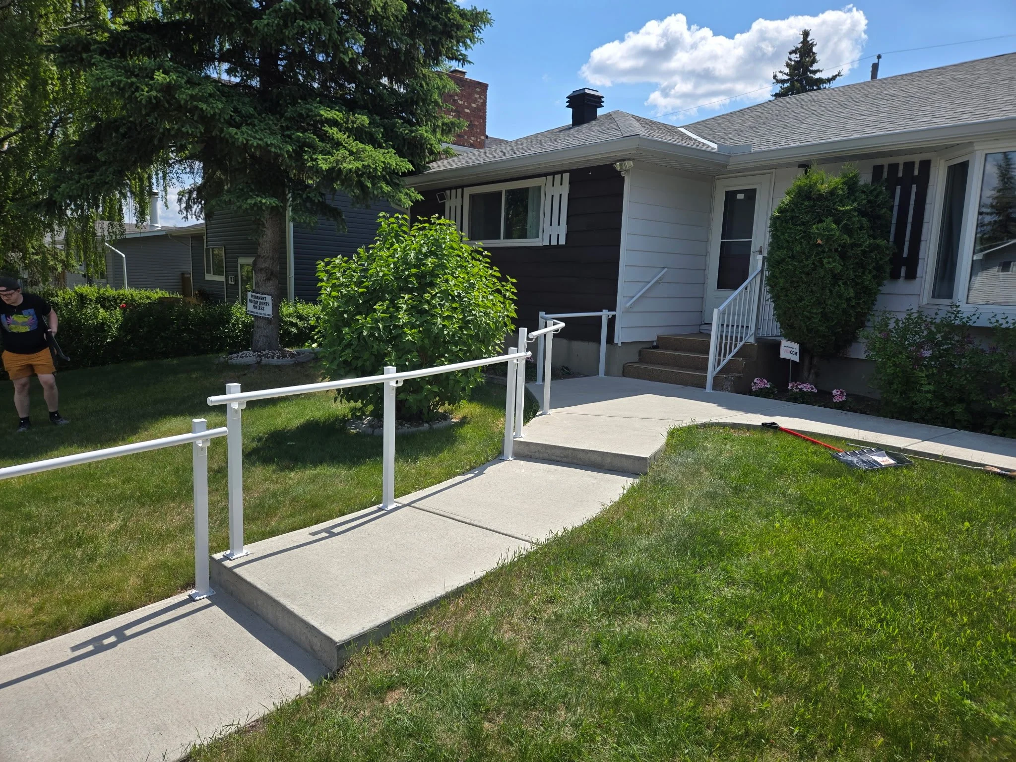 A house with a landscaped front yard featuring a small concrete ramp with white railings, a large evergreen tree, shrubs, and flowering plants. There is a person walking on the sidewalk.