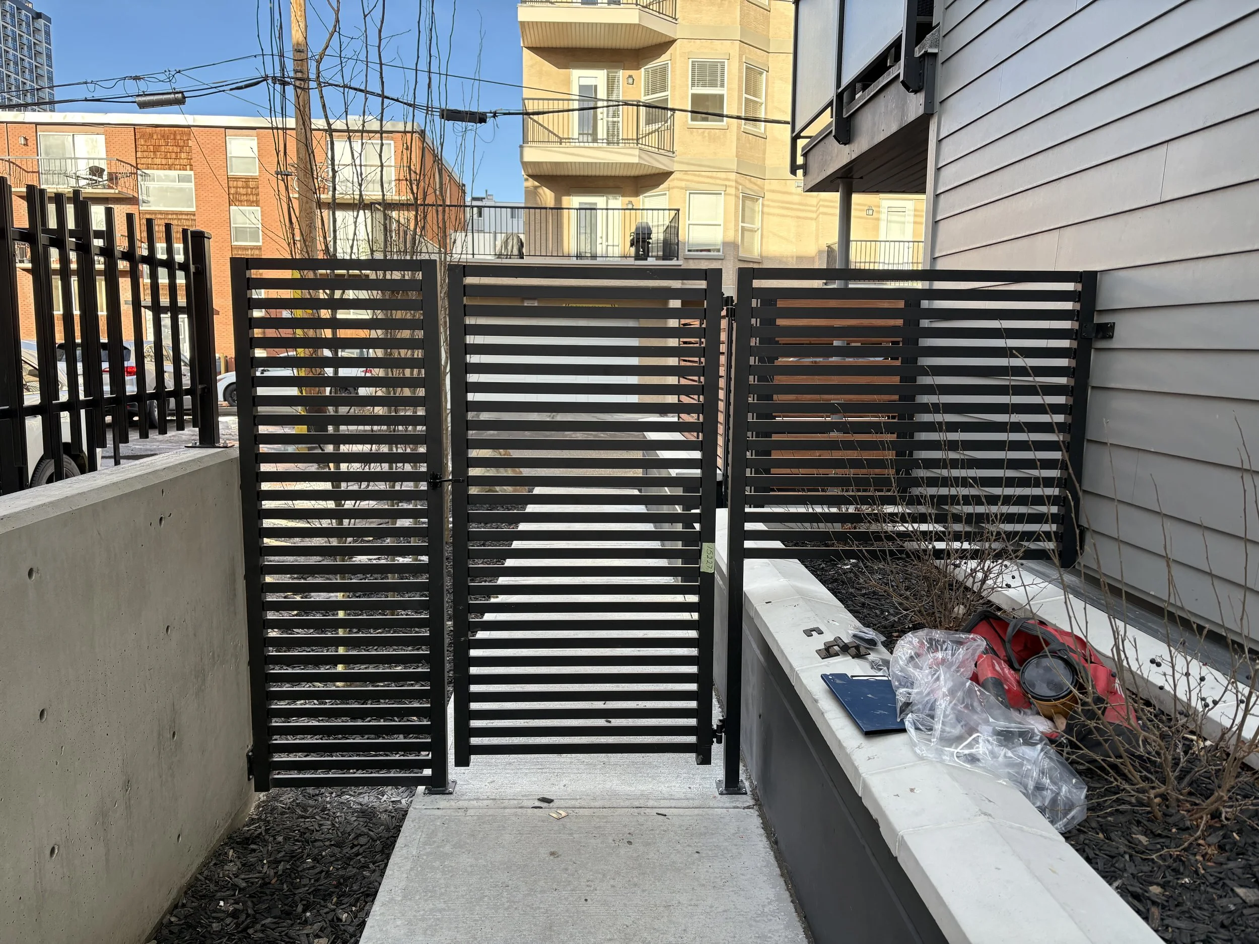 A modern balcony or patio area with a black metal gate, concrete flooring, and a planter filled with soil and small plants. In the background, there are neighboring multi-story buildings, some with brick facades and others with beige or gray siding. 