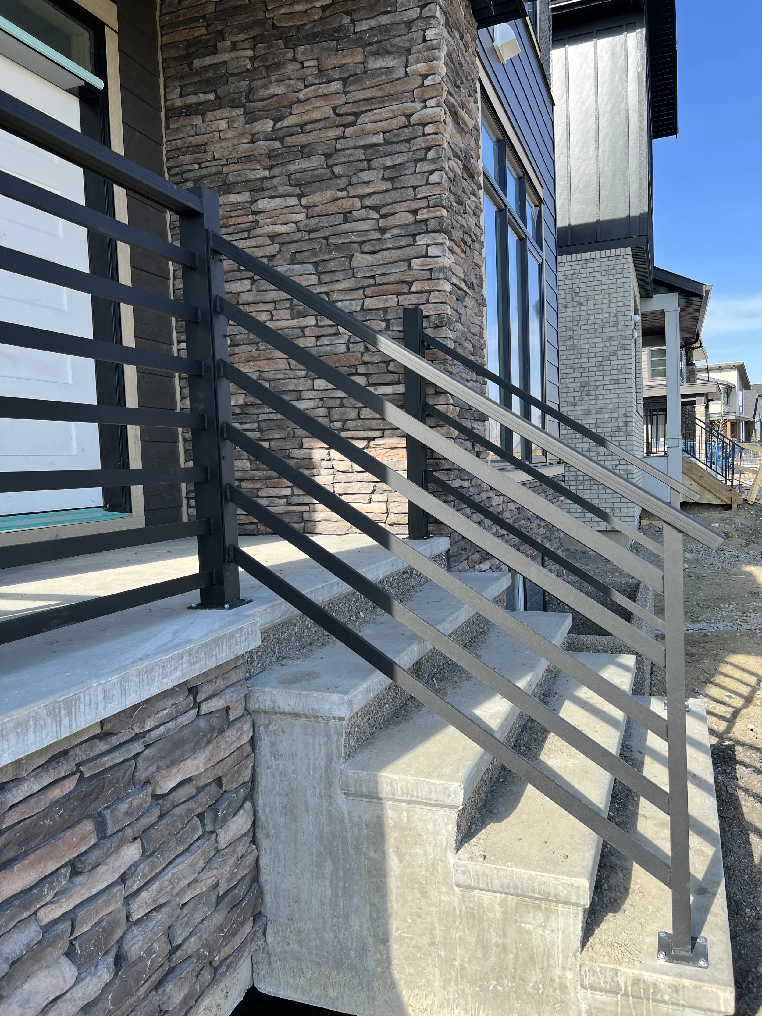 Exterior of a modern building with stone and brick walls, a large window, and a black metal staircase with a railing, leading up to the entrance. The building has multiple stories and a clear blue sky is visible.