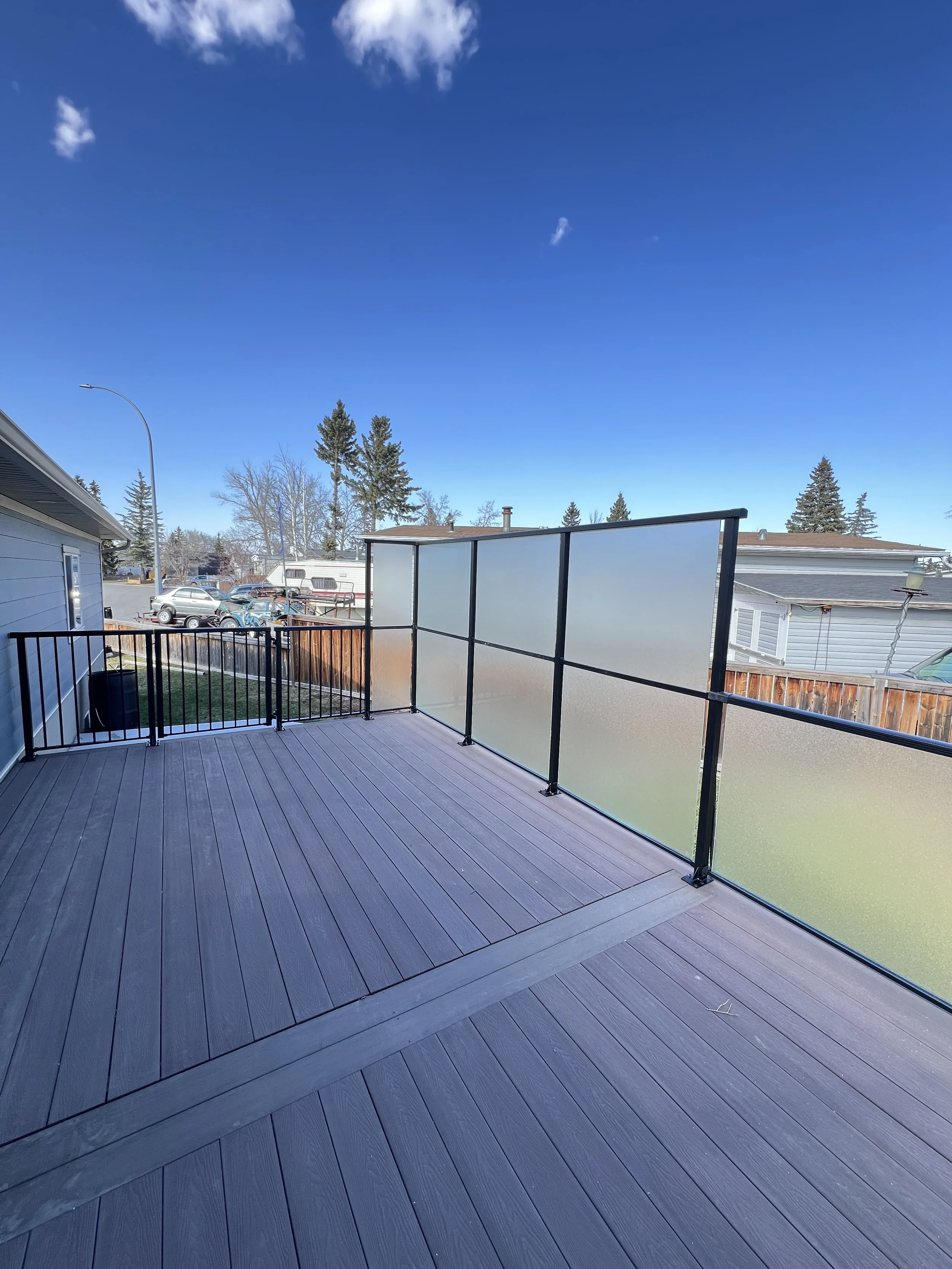 View from a balcony with dark wood decking, black metal railing, and frosted glass panels, overlooking a backyard with trees, cars, and neighboring houses under a blue sky with a few clouds.