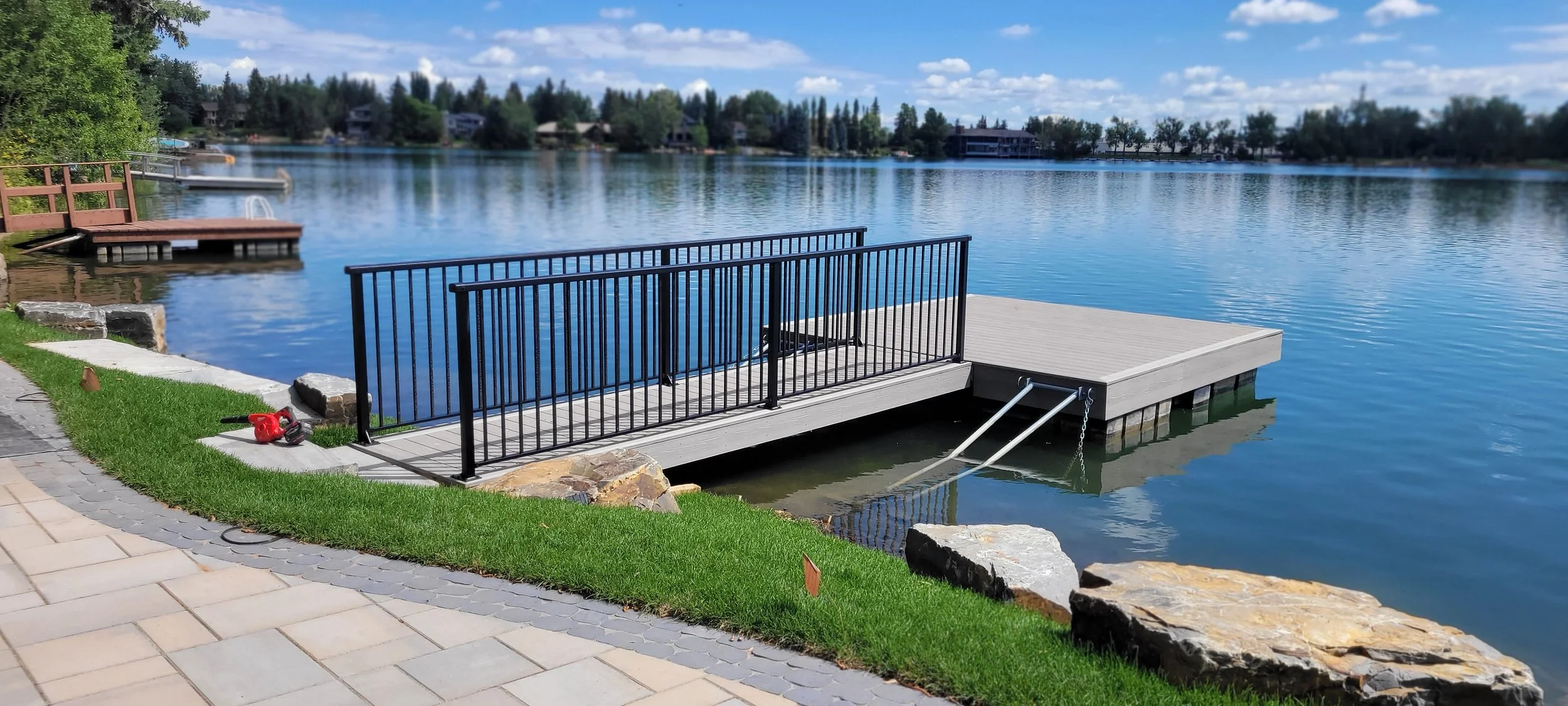 A lakeside dock with a black metal railing, extending over the water, surrounded by a grassy yard with paving stones and large rocks, under a partly cloudy sky.