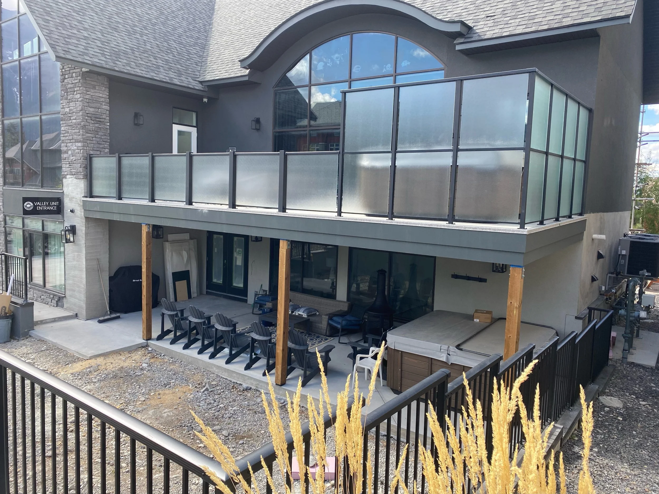 A modern multi-story building with a stone column, large glass windows, and a balcony with frosted glass panels. There is a patio with black chairs and a table, and a small fenced yard with some construction materials and gravel. Tall yellow grass is