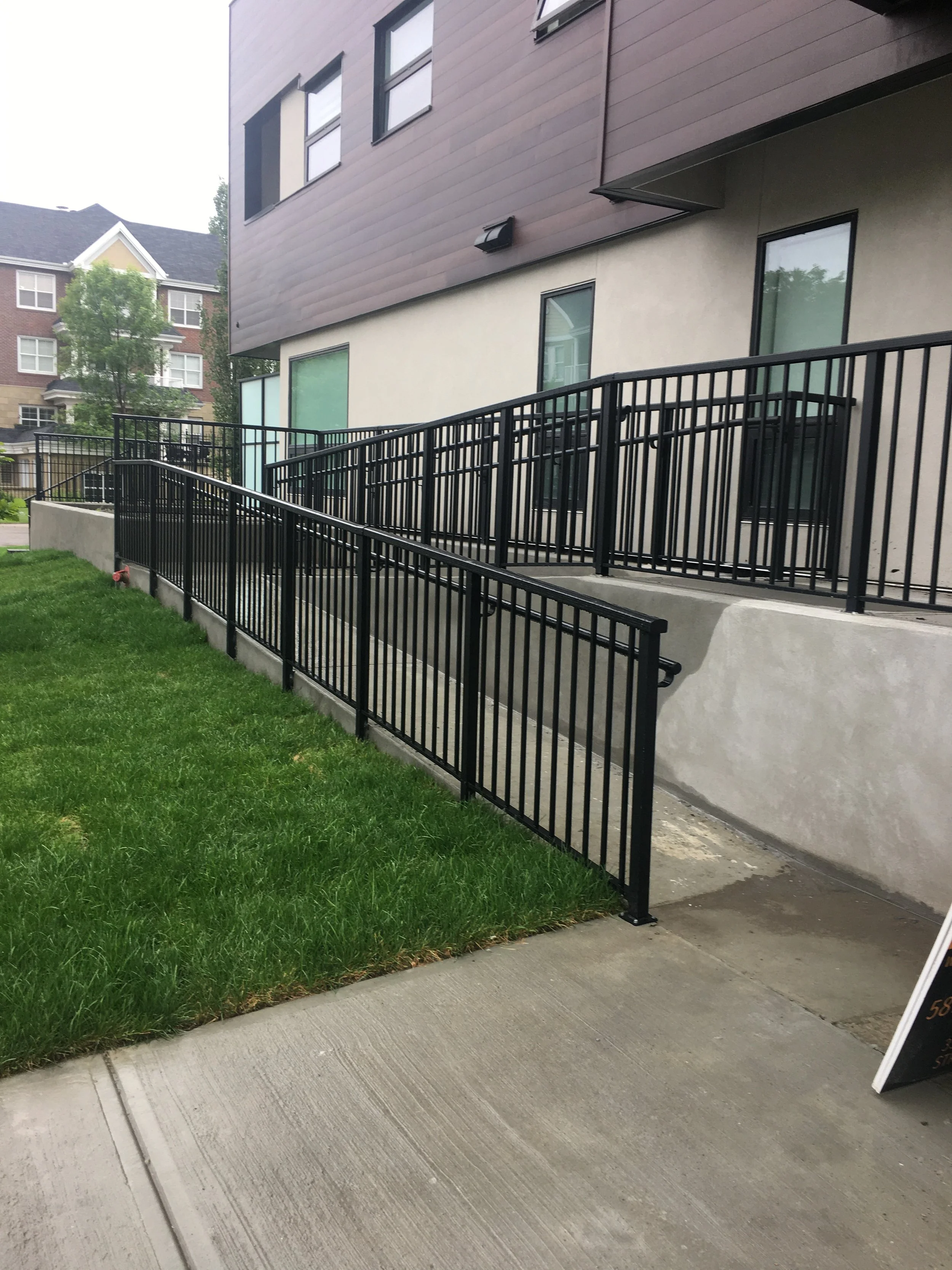A modern residential building with multiple windows and a black metal railing on a small staircase leading to the entrance. There is a grassy lawn and a sidewalk in front of the building, with some neighboring buildings visible in the background.