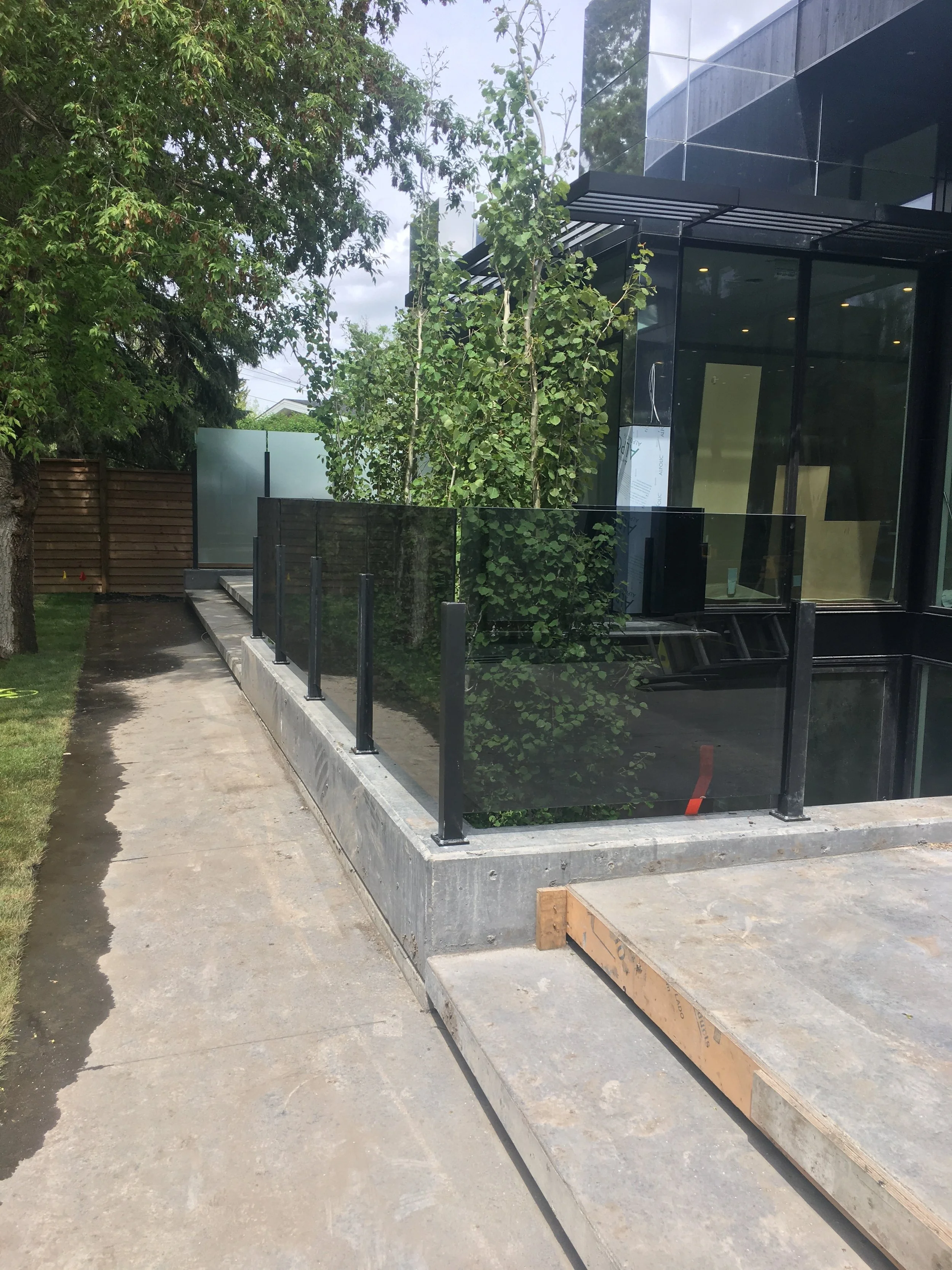 Construction site with a modern glass building, small trees, and a concrete sidewalk with a metal fence.