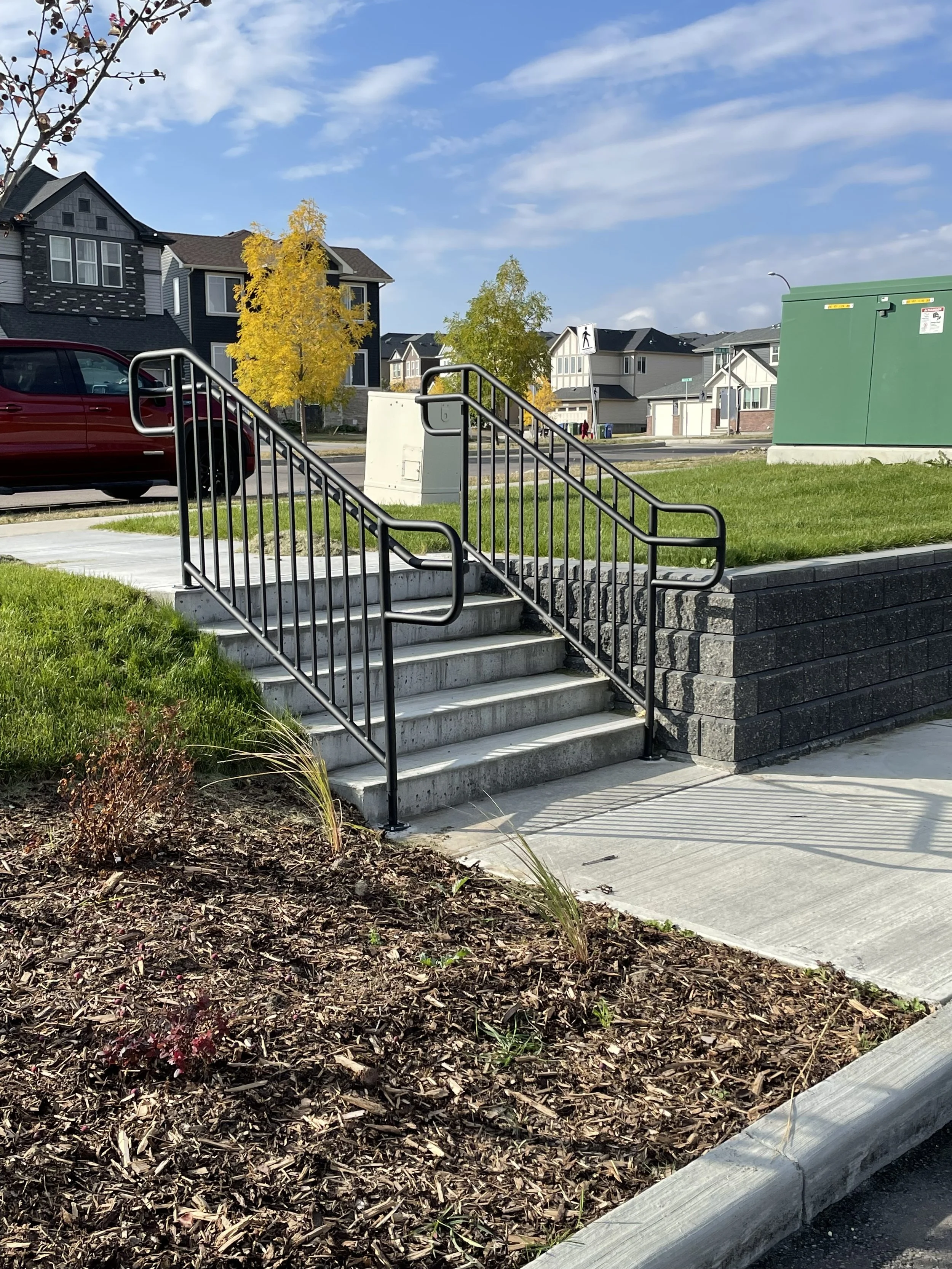 Residential neighborhood sidewalk with stairs, metal handrails, green utility box, and a flower bed in the foreground, with houses and trees with yellow and green leaves under a blue sky with clouds.