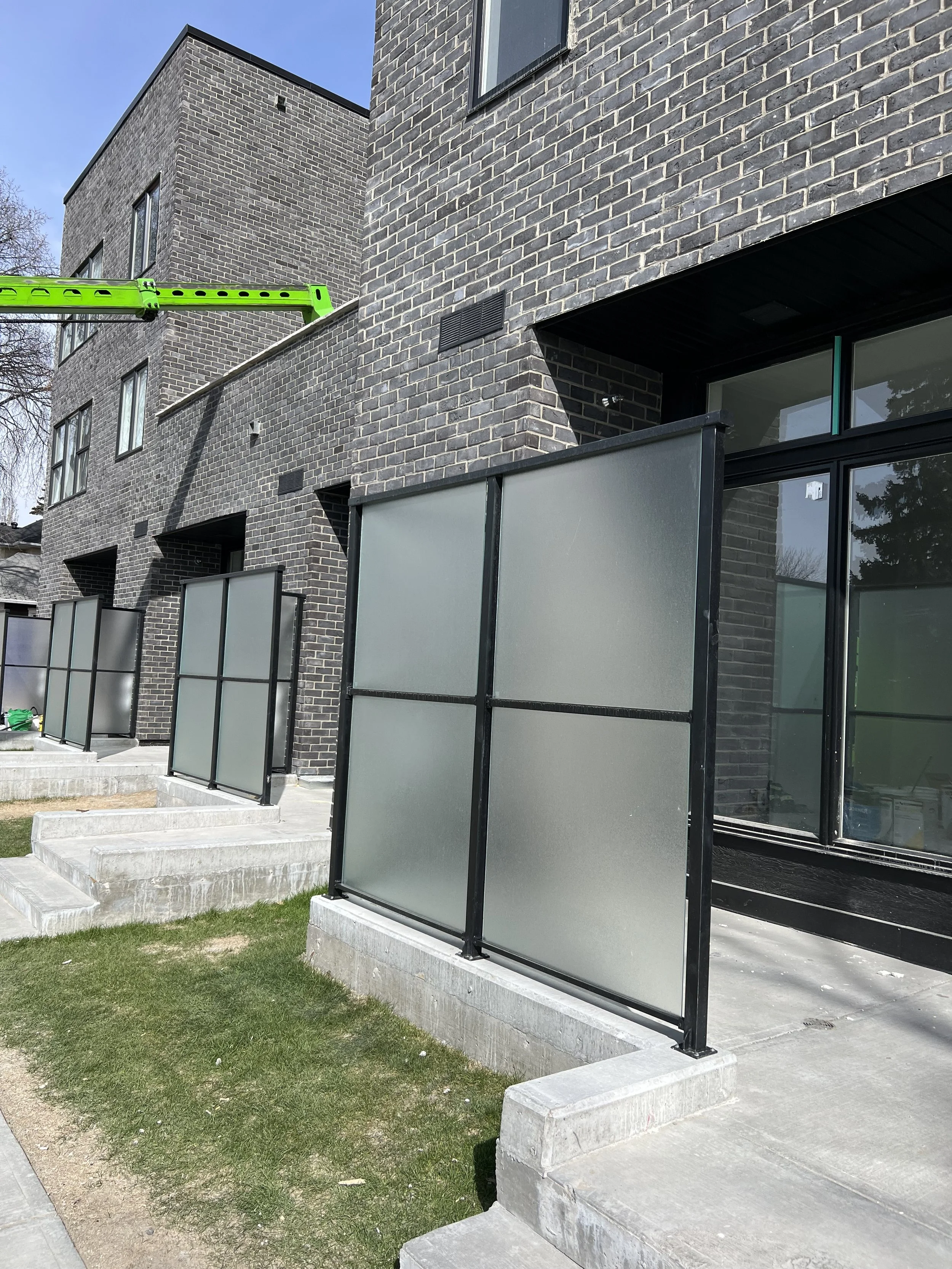 Modern apartment building with black brick exterior, glass doors with black frames, and frosted glass privacy panels on the balconies. Concrete steps lead up to the entrances, with a small grass patch and sidewalk in front.