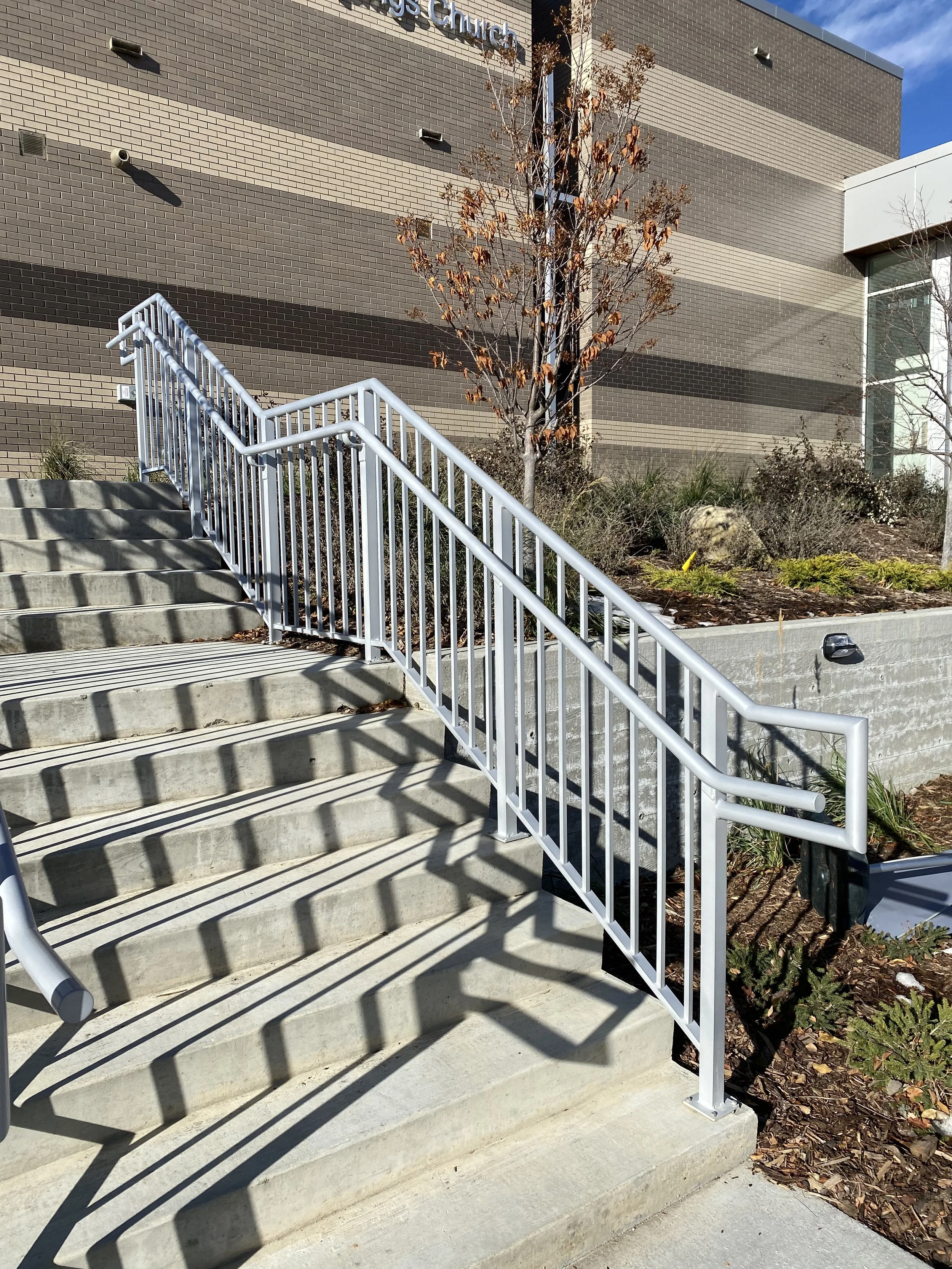 Concrete stairs with a white metal railing leading up to a brick building with trees and shrubs beside it.