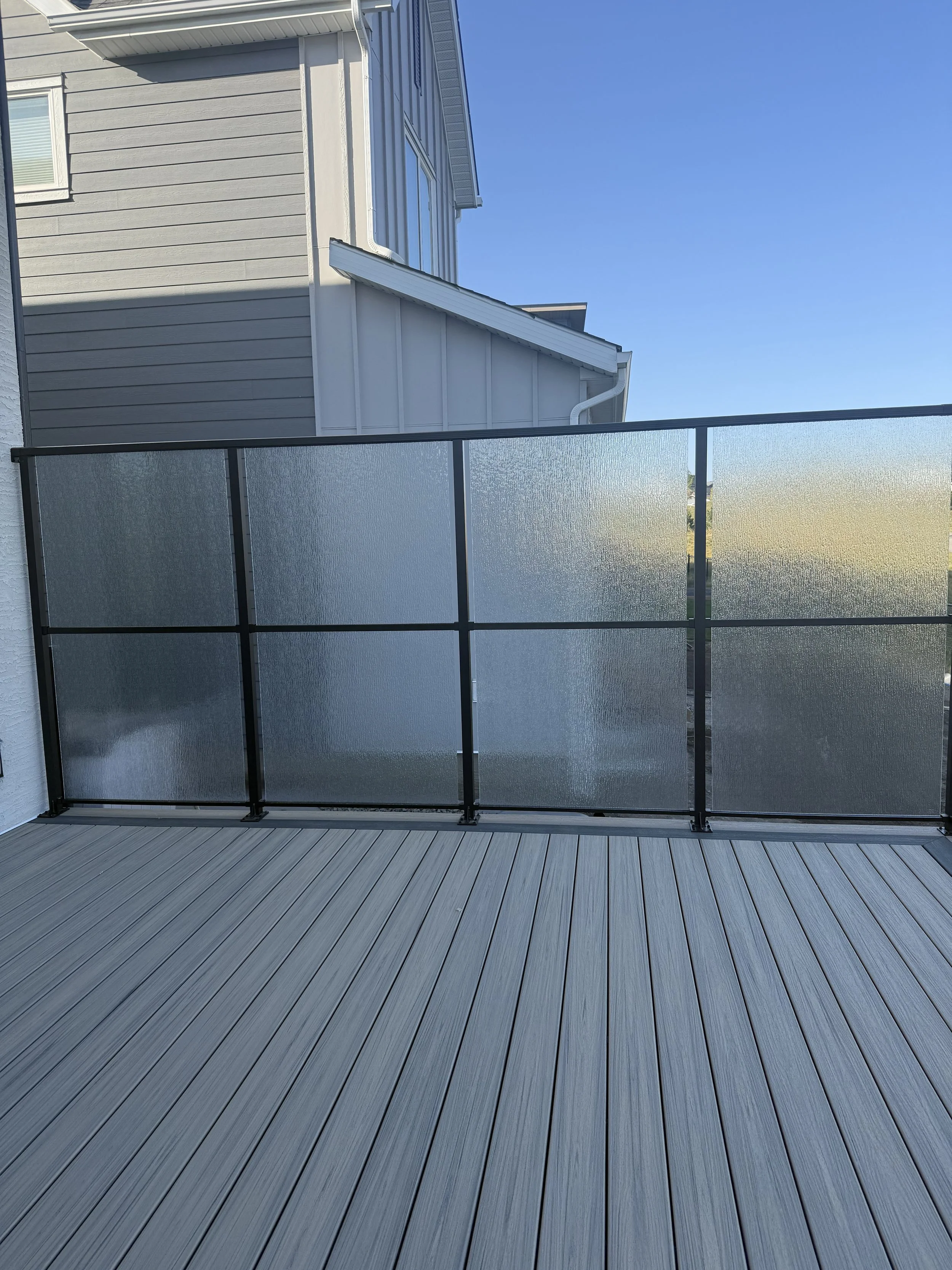 A patio or balcony with grey wooden decking and frosted glass panels on black metal frames, with a neighboring house showing grey siding and a white roof in the background under a clear blue sky.
