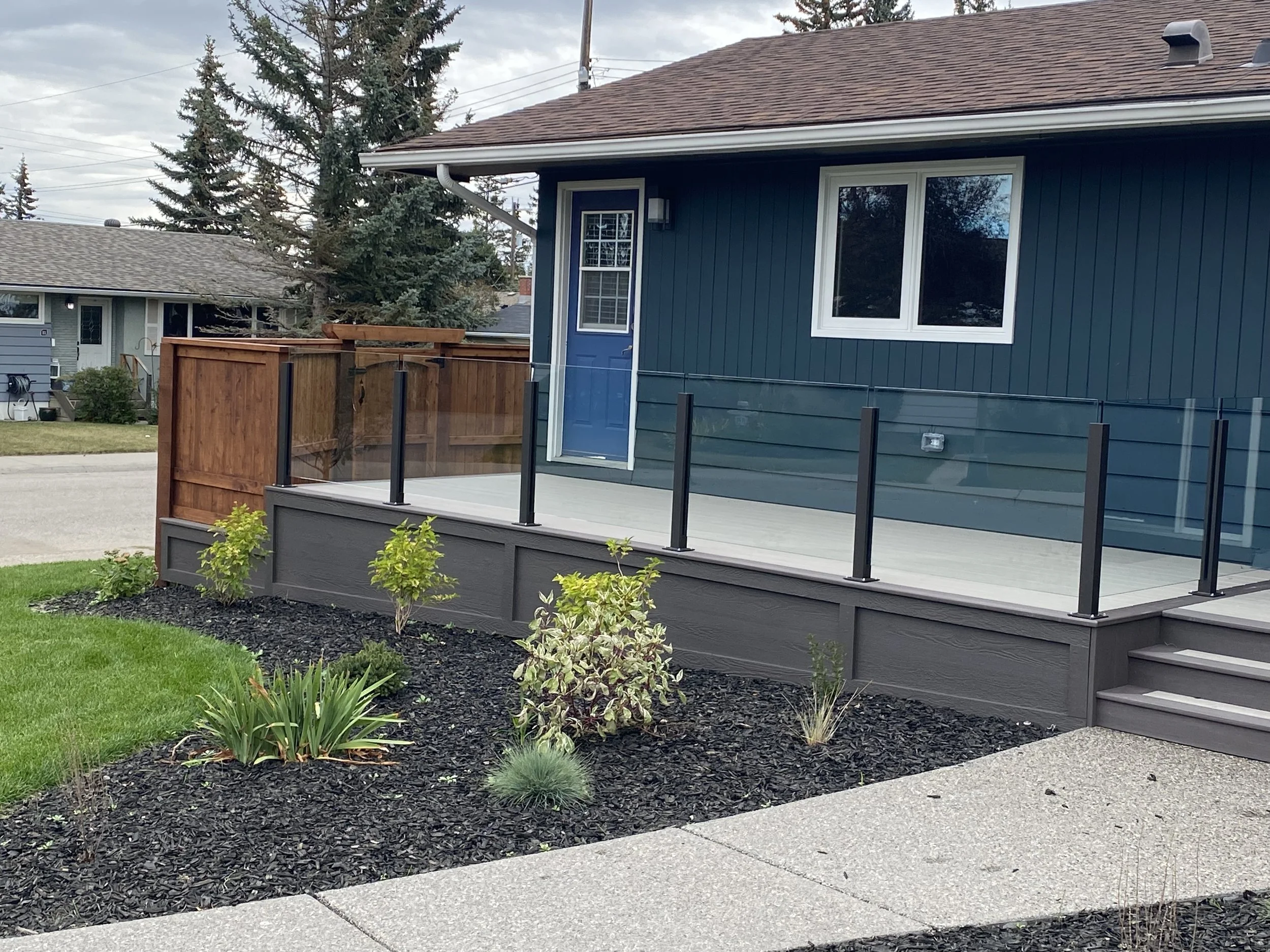 A house with a blue exterior and white trim features a small front deck with black railings and stairs. There are plants and shrubs in a flower bed with dark mulch in the front yard. The house has a large window and a smaller window next to a blue do