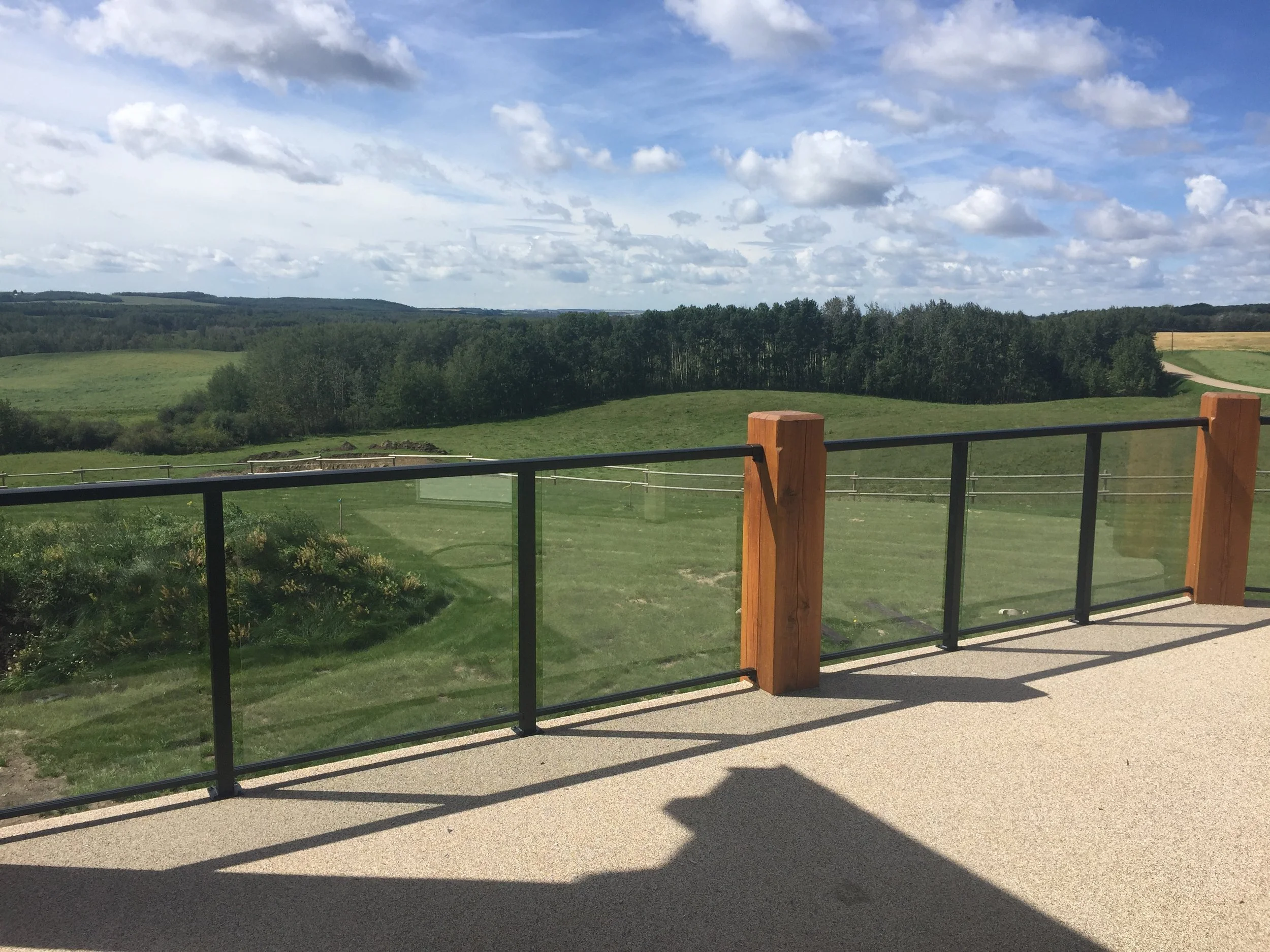 View of green fields and trees under a partly cloudy sky, seen from a balcony with glass and wooden railing.