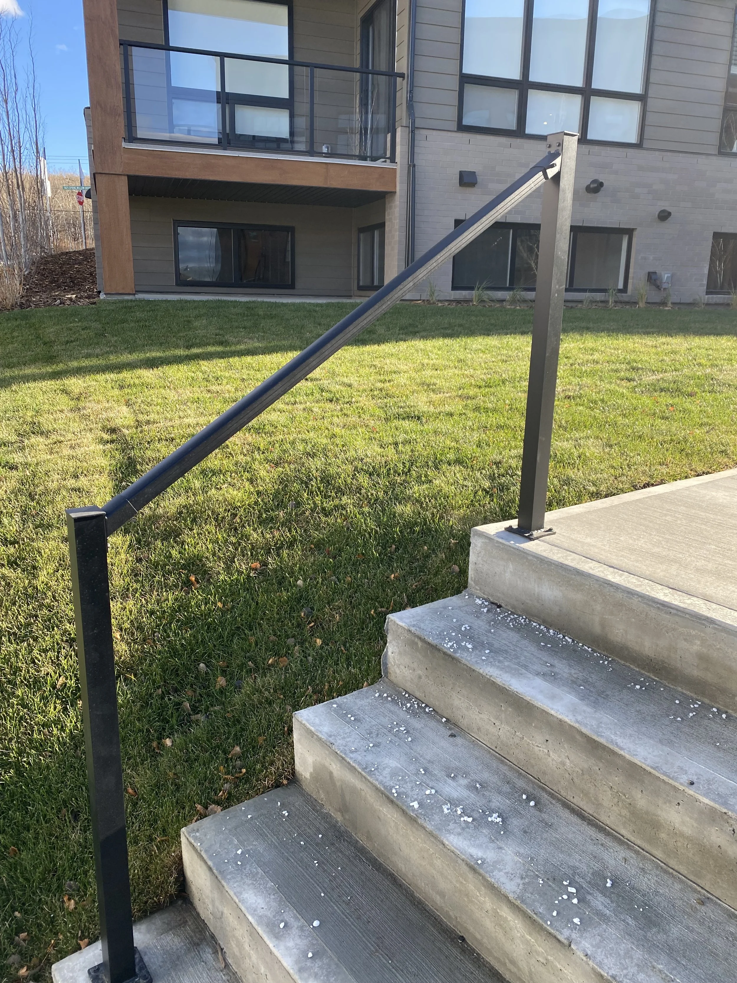 Concrete stairs with black metal handrail leading to a building entrance, with a grassy lawn on the side and a modern apartment complex in the background.