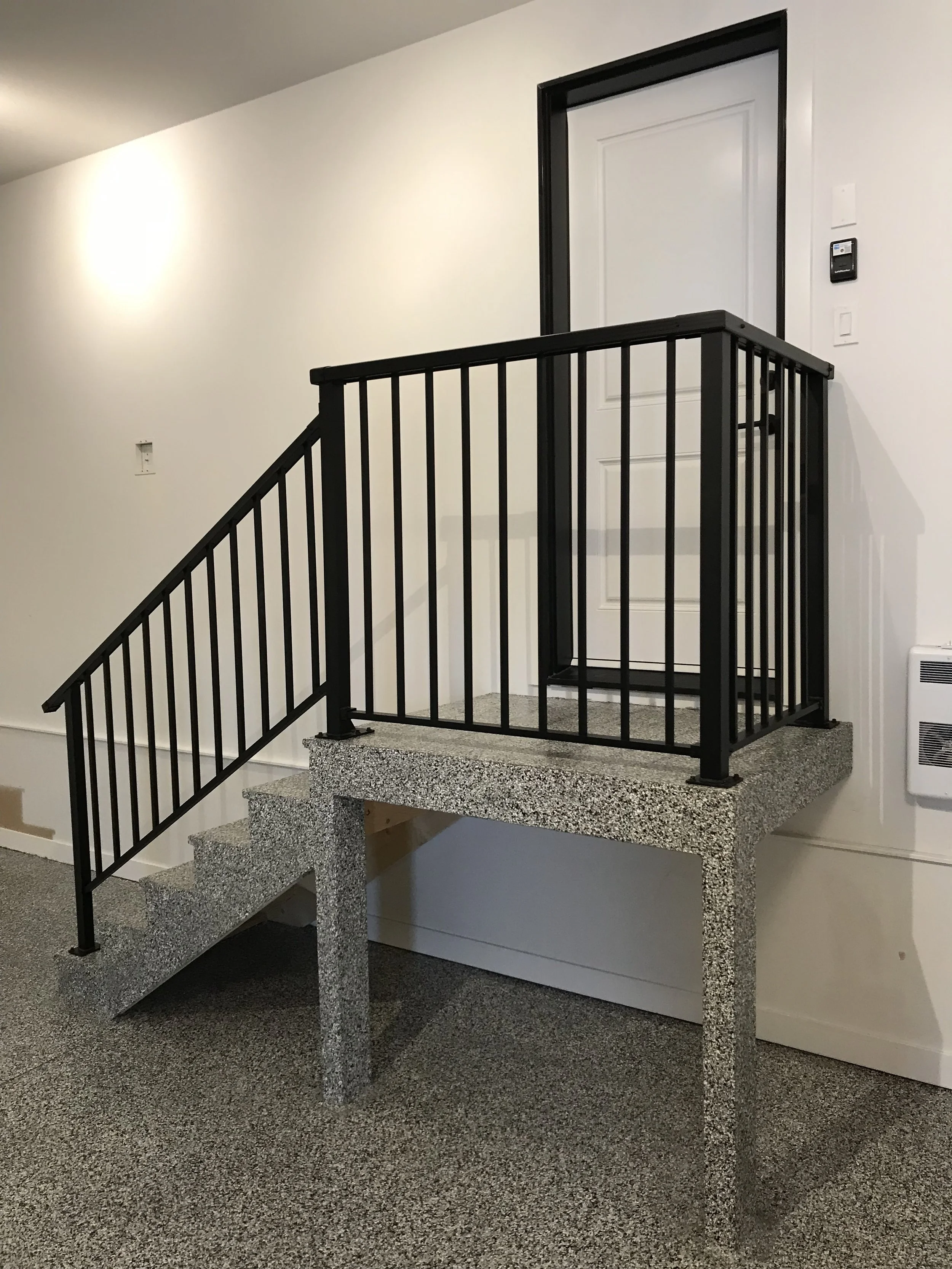 Interior of a building with stairs made of speckled gray concrete, metal black railings, and a door on the upper level, with white walls and carpeted flooring.
