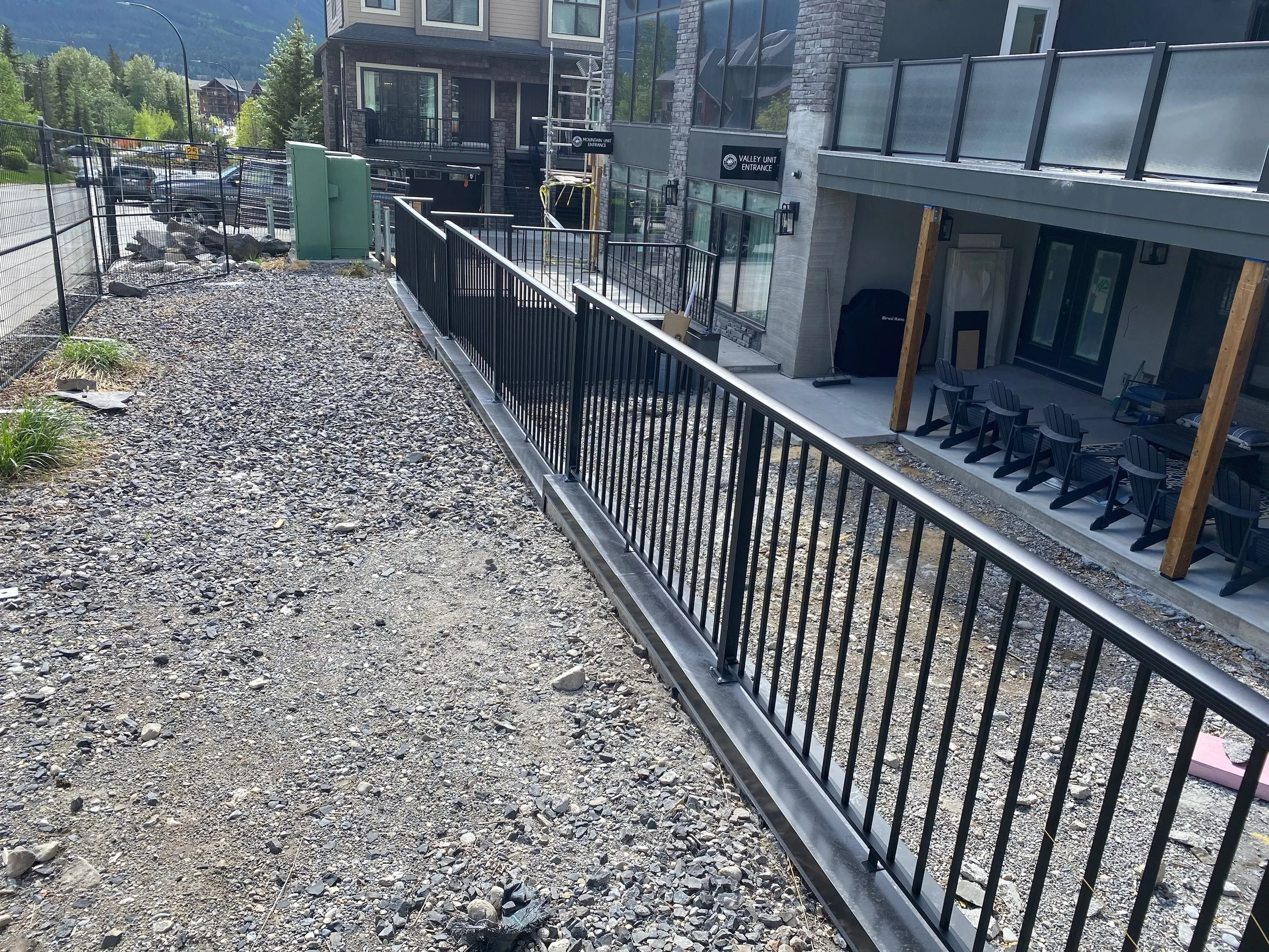 Construction site showing a building with large glass windows, a metal railing and a gravel ground. Signs indicate the entrances for 'Valley Unit' and 'Mountain Unit'. There are chairs, some tools, and unfinished wooden posts on the patio area.