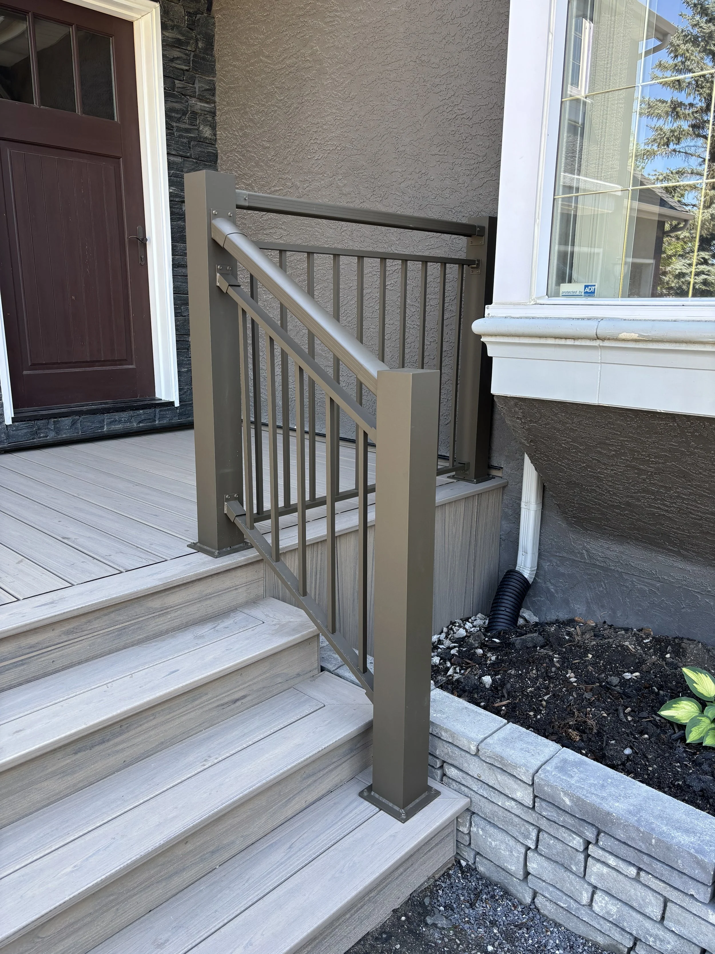 Exterior of a house with light-colored wooden stairs and a brown front door. There is a gray metal handrail on the stairs, a stone accent wall adjacent to the door, and a large window with a white frame on the right side. Part of a black drainage pip