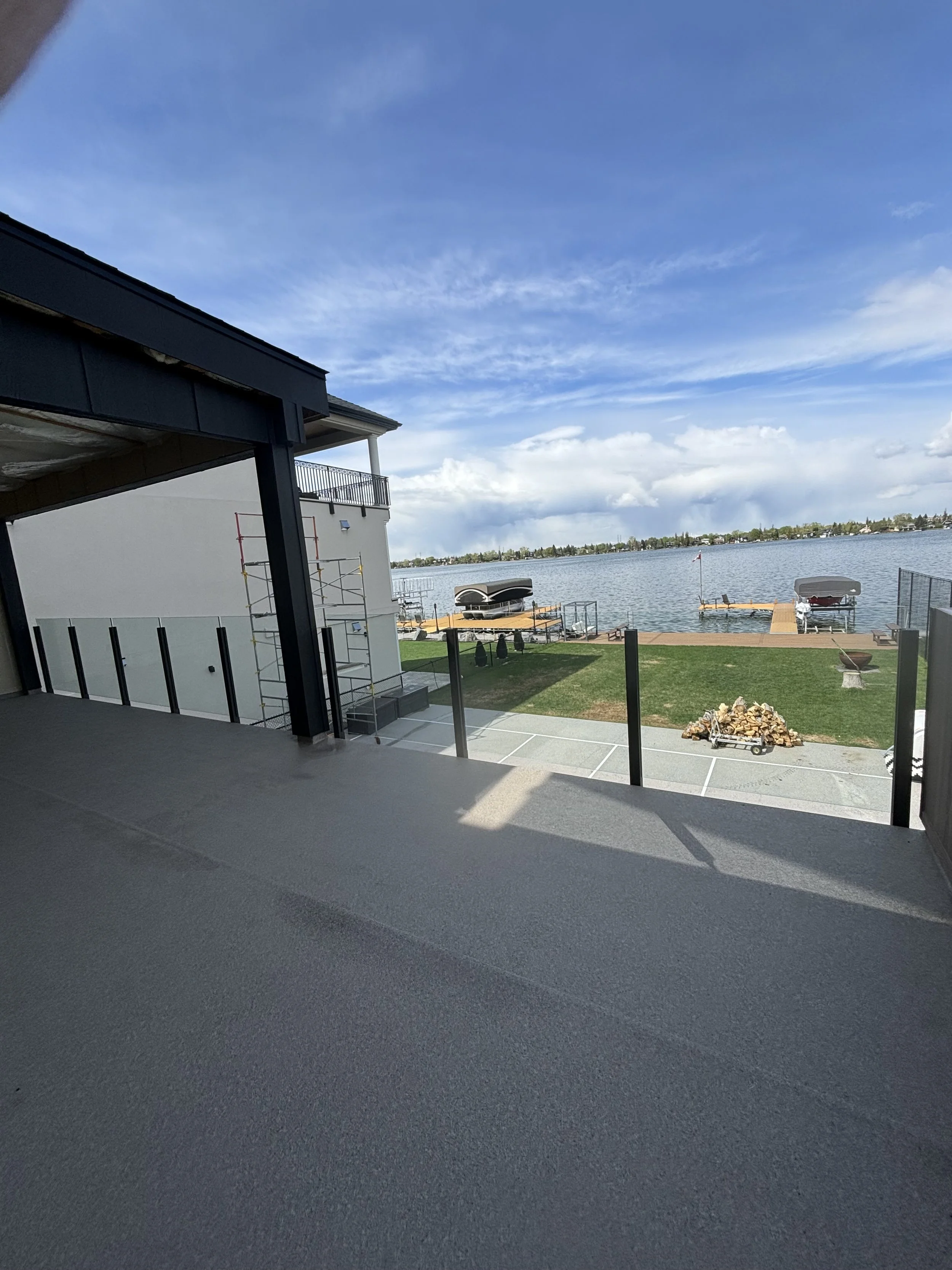 View of a lakeside scene seen from a covered patio with a black railing. The scene includes a dock with boat lifts, a floating boat, and a distant shoreline with houses and trees under a partly cloudy sky.