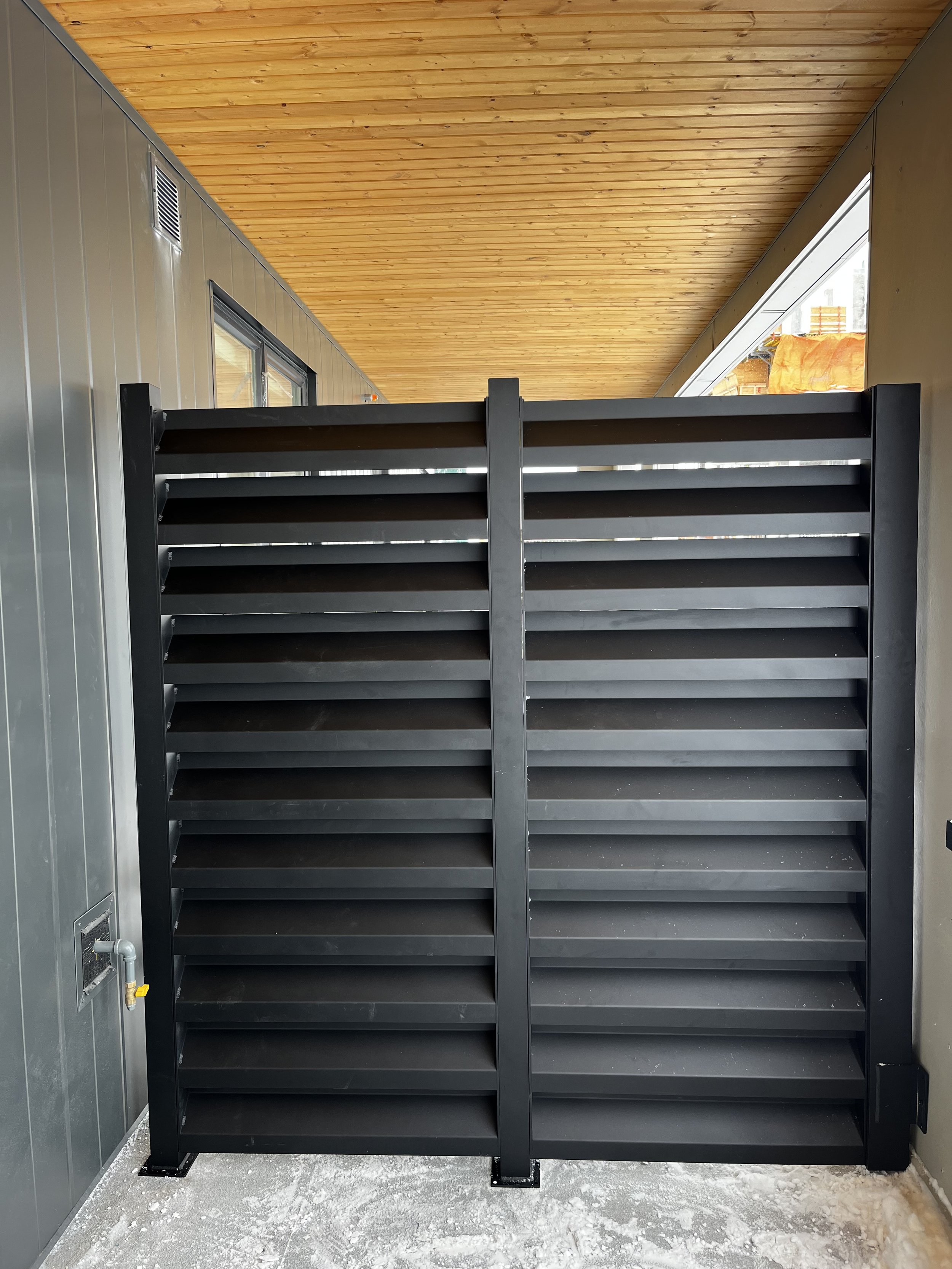 Black outdoor storage rack with multiple shelves, placed in front of a gray wall with a window, beneath a wooden ceiling, with snow on the ground.