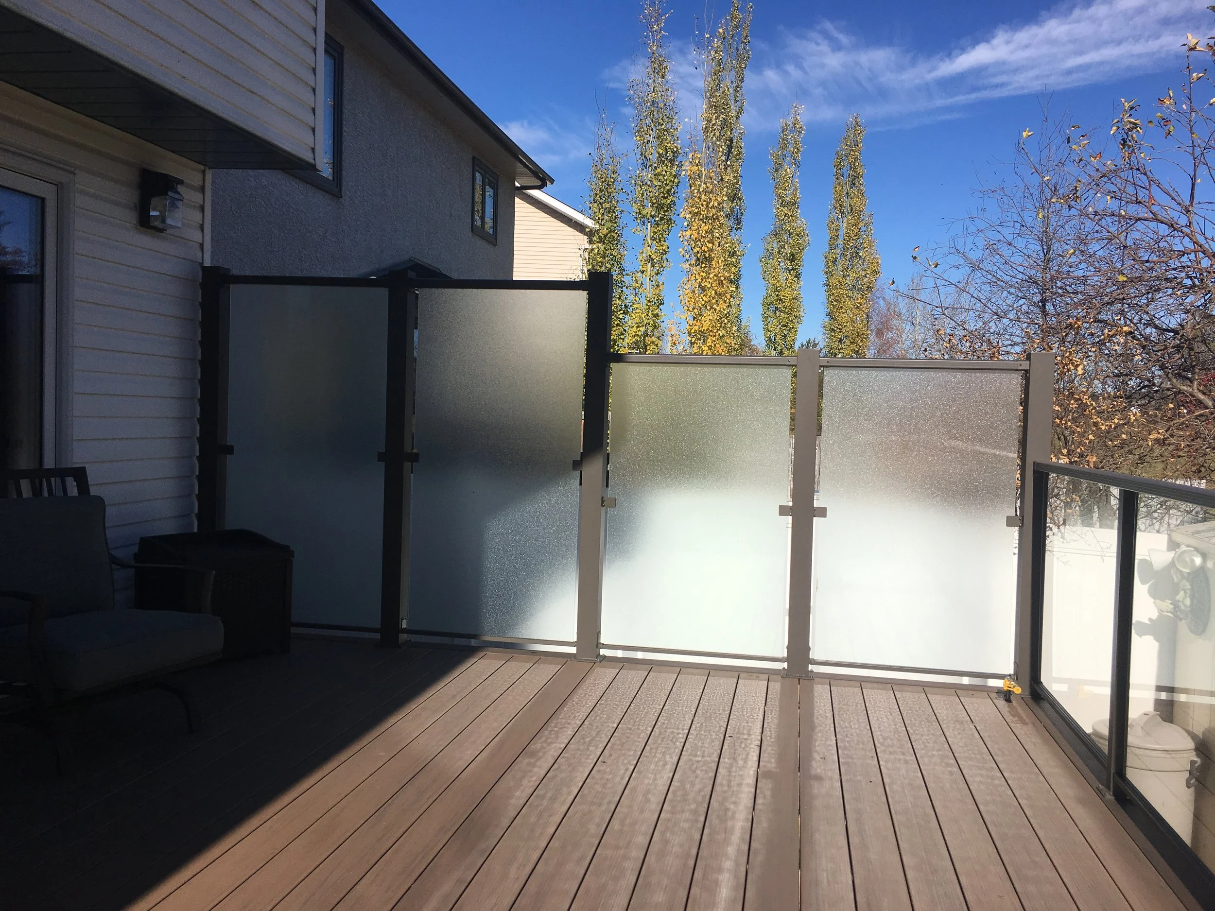 A balcony with wooden flooring and frosted glass panels, with a patio chair in the lower left corner, overlooking trees with yellow and green leaves under a blue sky.