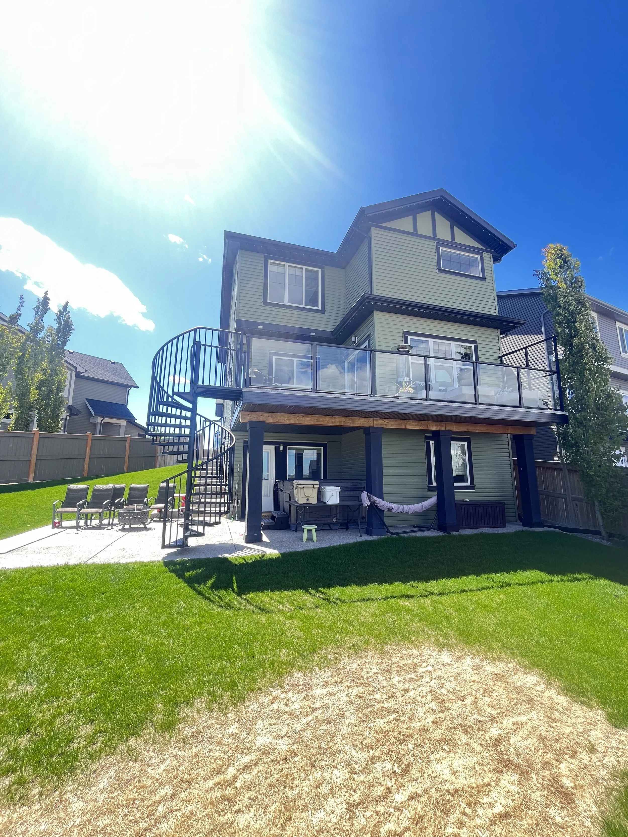 A multi-story house with a green exterior, black trim, and a large balcony with a glass railing, set in a backyard with a lawn and a gravel pathway under bright sunlight.