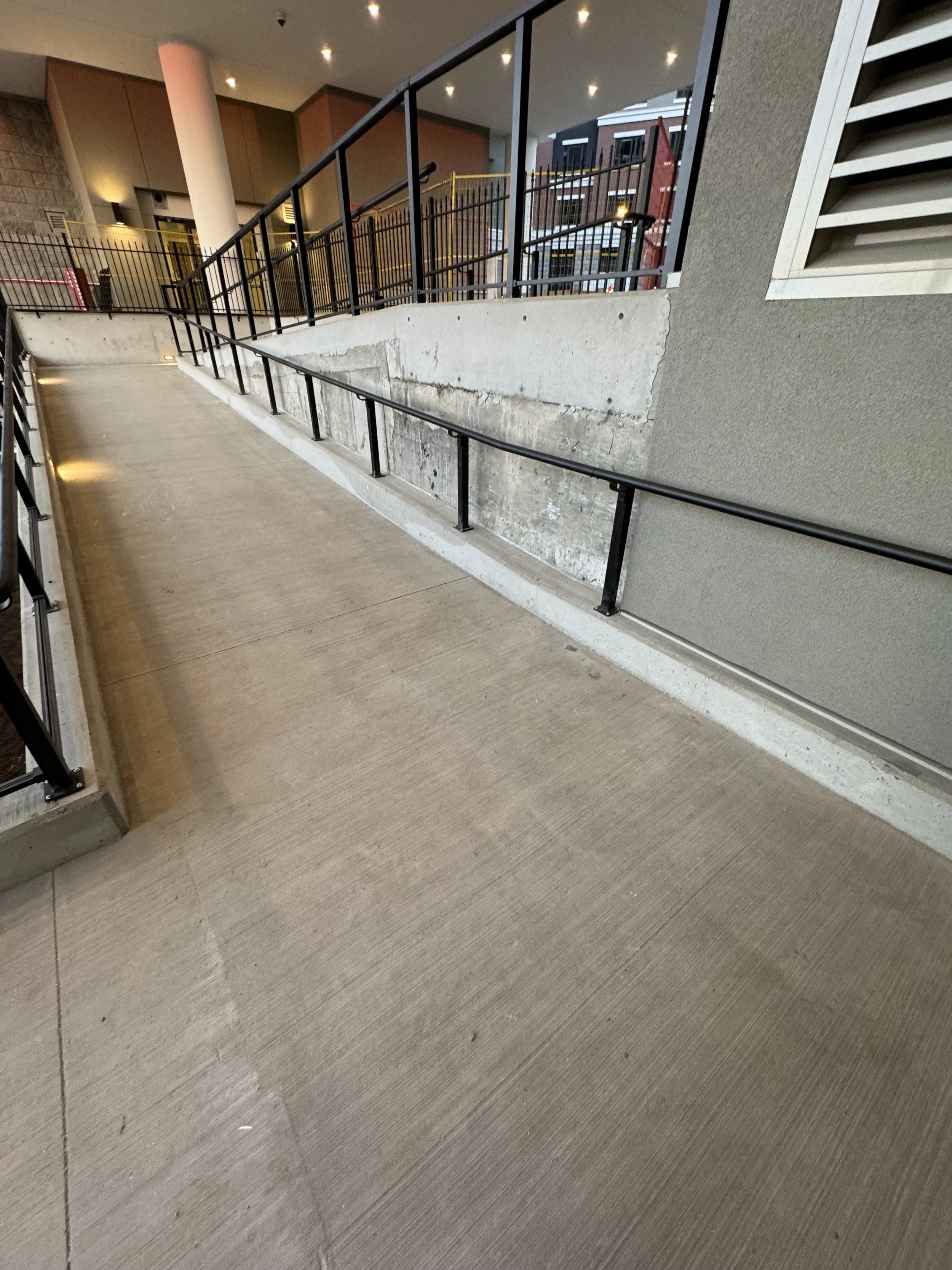 Interior of a modern building with a sloped wheelchair-accessible ramp, concrete and beige walls, black metal railings, and a multi-level stairway on the right side. Buildings with windows are visible outside.