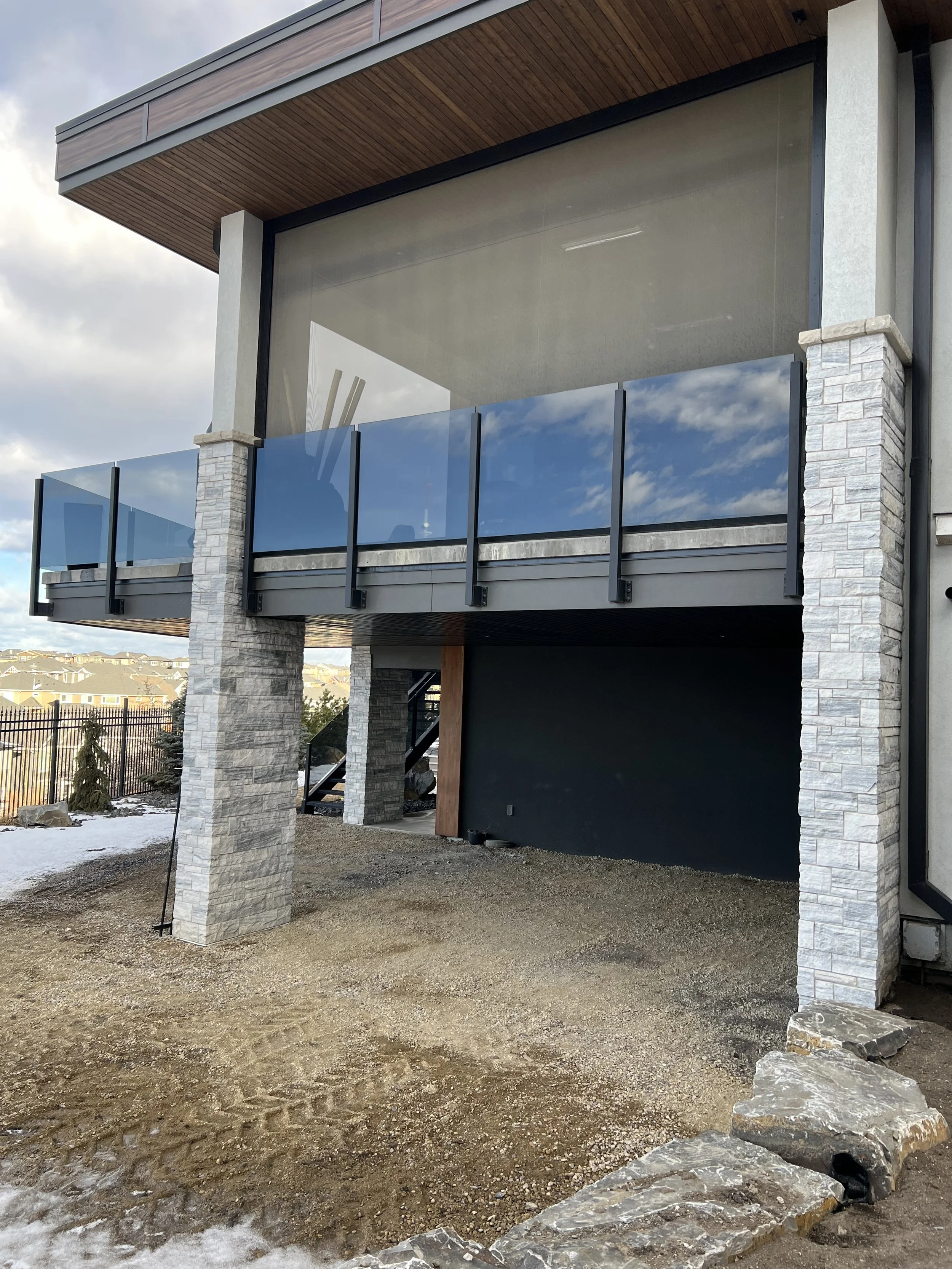 Exterior of a modern building with large glass windows, stone columns, and a wooden overhang. The ground is dirt with some snow and rocks.